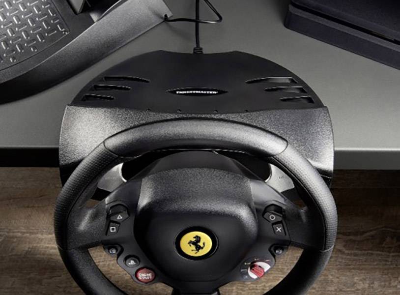 Un volante nero con il logo Ferrari, montato su una scrivania. Sopra di esso ci sono i pedali e una console per videogiochi come accessori.