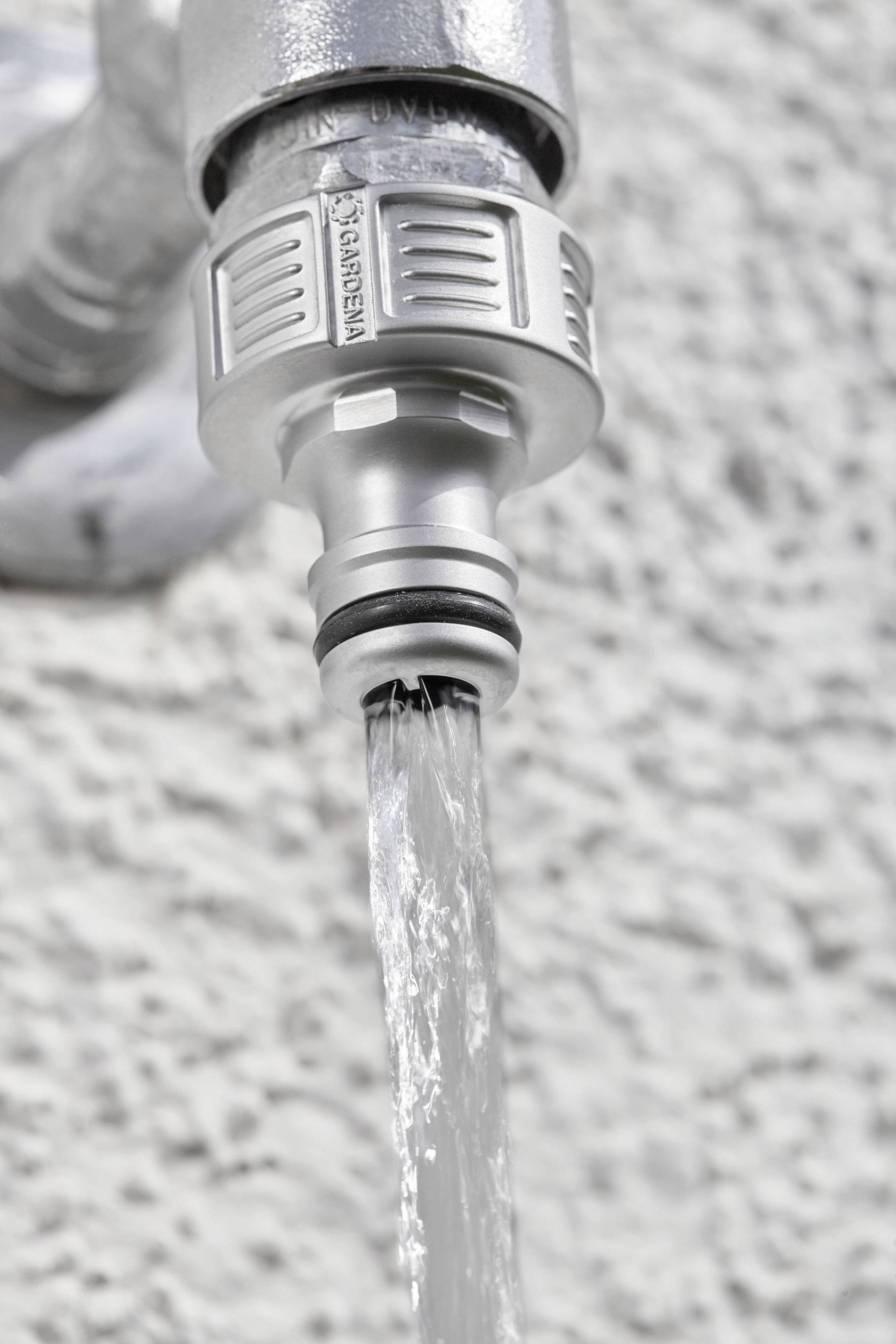 L'acqua scorre da un rubinetto argentato su una parete grigia. Il focus è sul getto d'acqua e sul punto di connessione del rubinetto.
