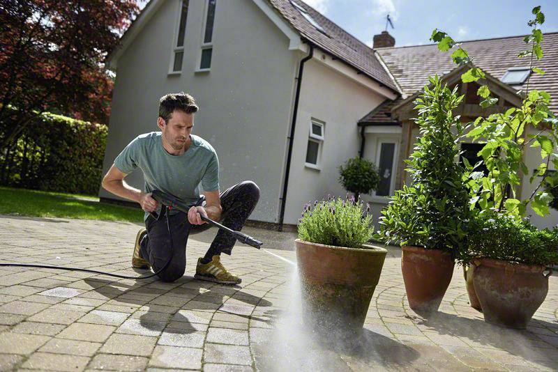 Una persona sta pulendo la terrazza in giardino con un idropulitrice. Sullo sfondo si vedono piante in vasi e una casa.