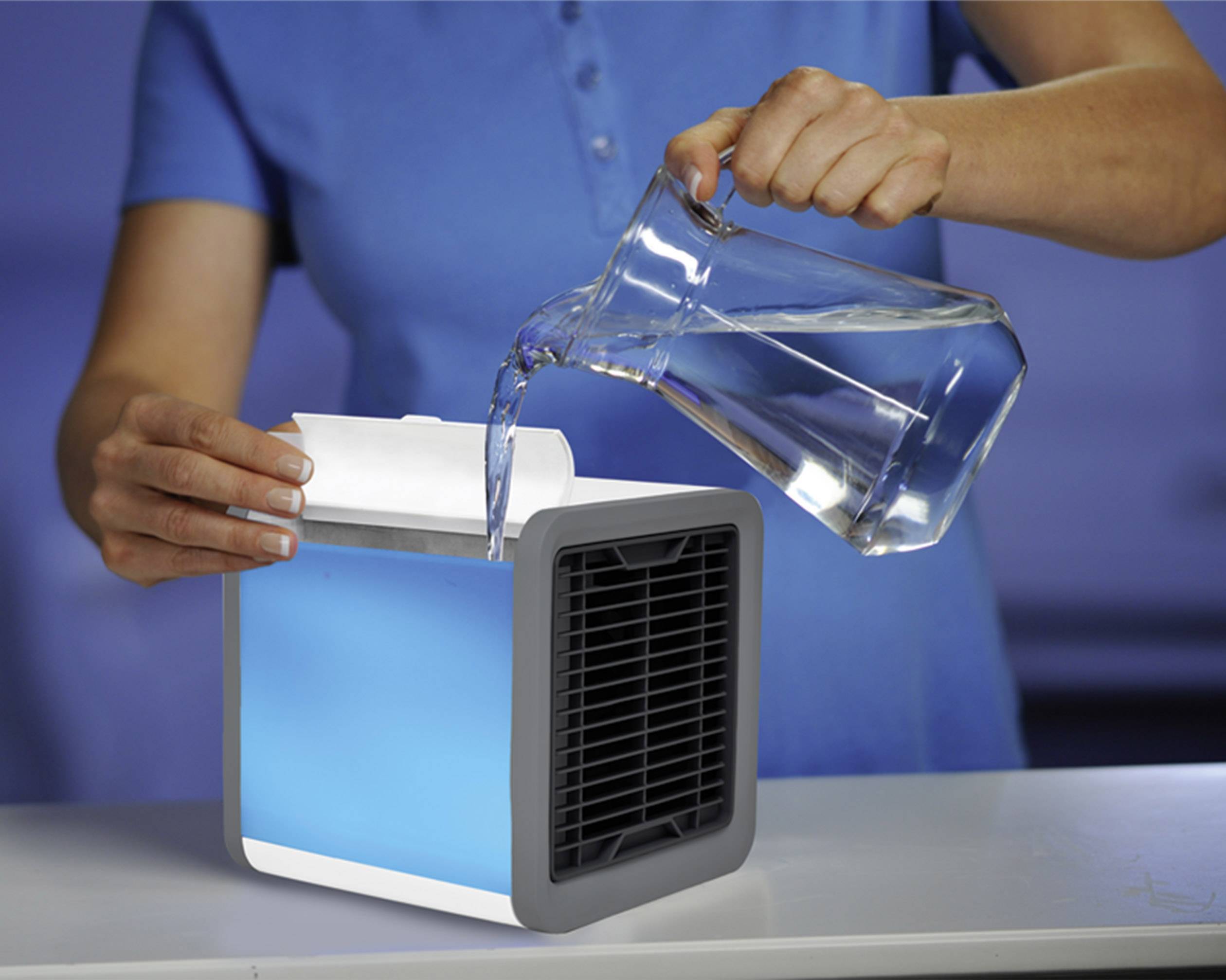 Una persona versa acqua in un piccolo condizionatore portatile blu posizionato su un tavolo. Il condizionatore ha una griglia anteriore nera.
