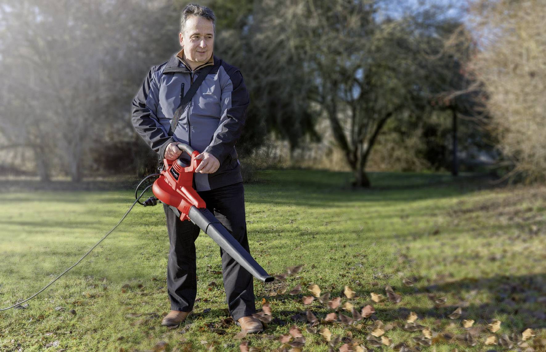 Un uomo con una giacca scura sta utilizzando un soffiatore rosso per rimuovere le foglie autunnali da un prato verde.