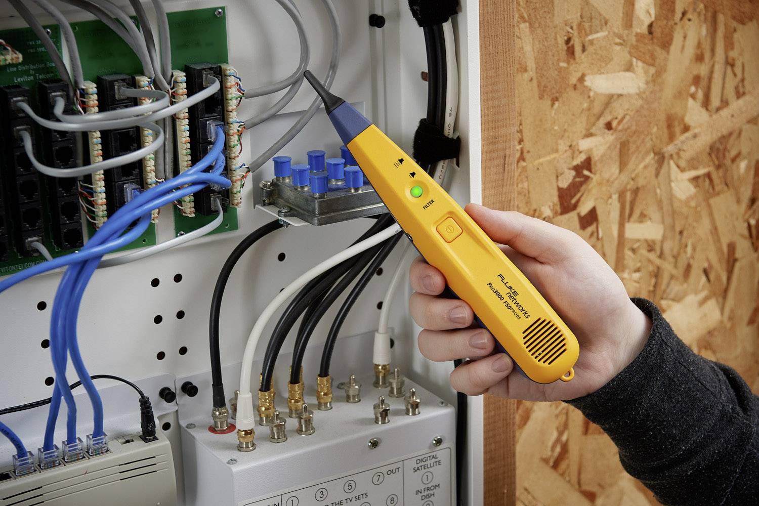 Una mano tiene un tester giallo che viene utilizzato su un quadro elettrico aperto con cavi e connessioni.