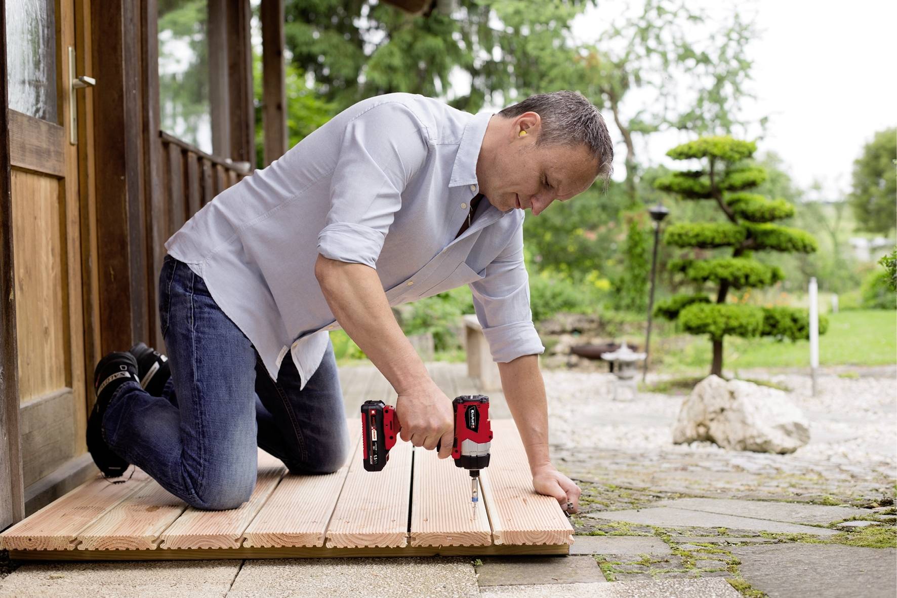 Un uomo è inginocchiato su una terrazza in legno e sta fissando delle tavole con un avvitatore a batteria. Sullo sfondo un giardino curato.