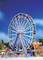 Una grande ruota panoramica in una fiera sotto un cielo blu. Persone passeggiano intorno. Attrazioni colorate e bancarelle sullo sfondo.