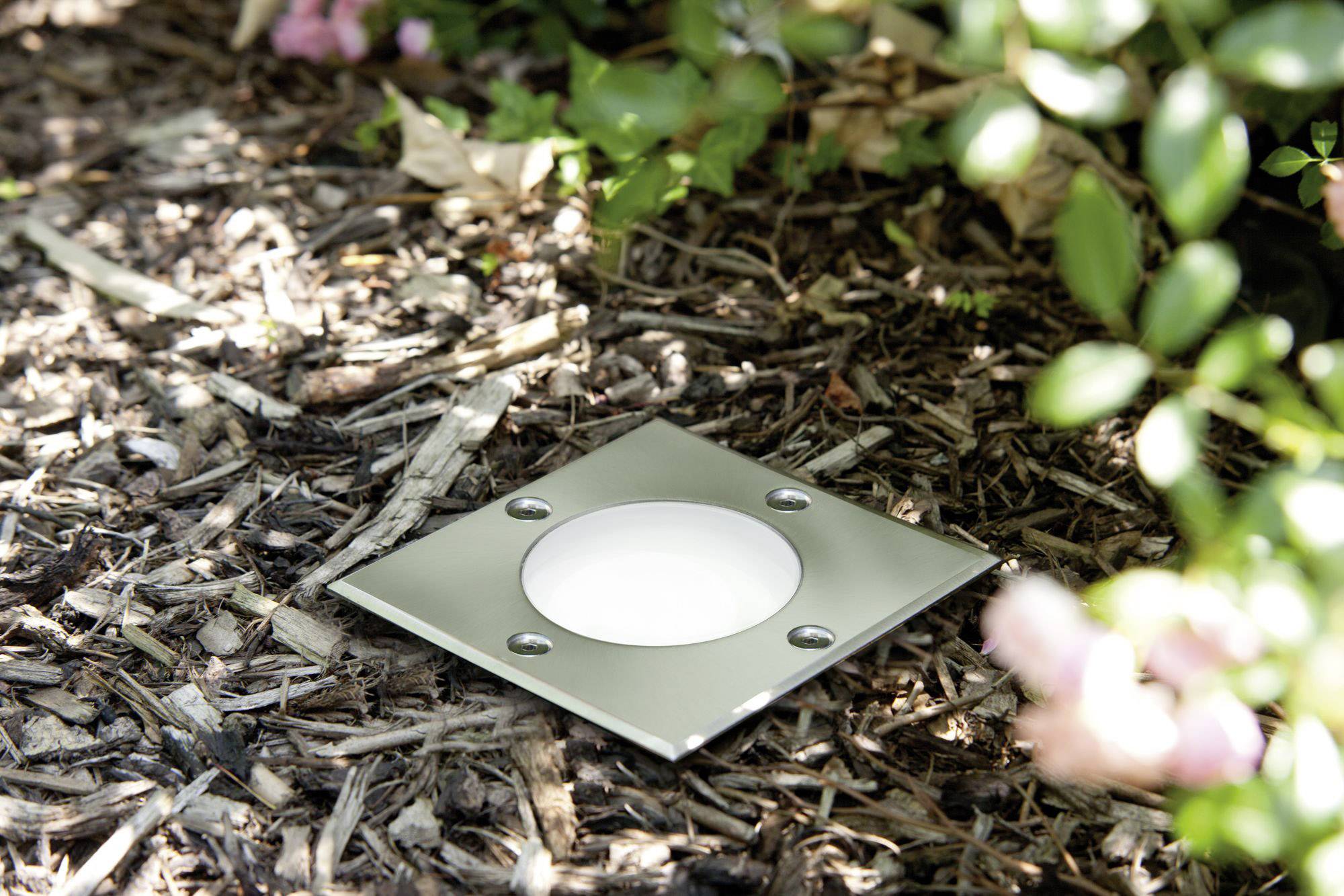 Lampada da incasso a pavimento con cornice quadrata in metallo in un giardino, circondato da pacciame e piante. Illuminazione per esterni.