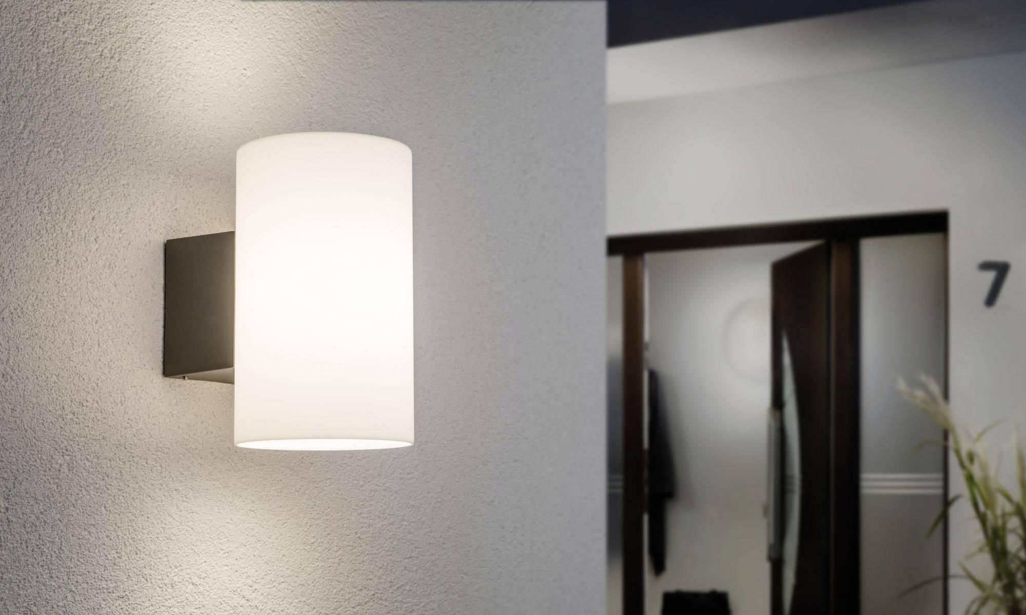 Una moderna applique con paralume cilindrico bianco illumina un corridoio. Sullo sfondo è visibile una porta leggermente socchiusa.