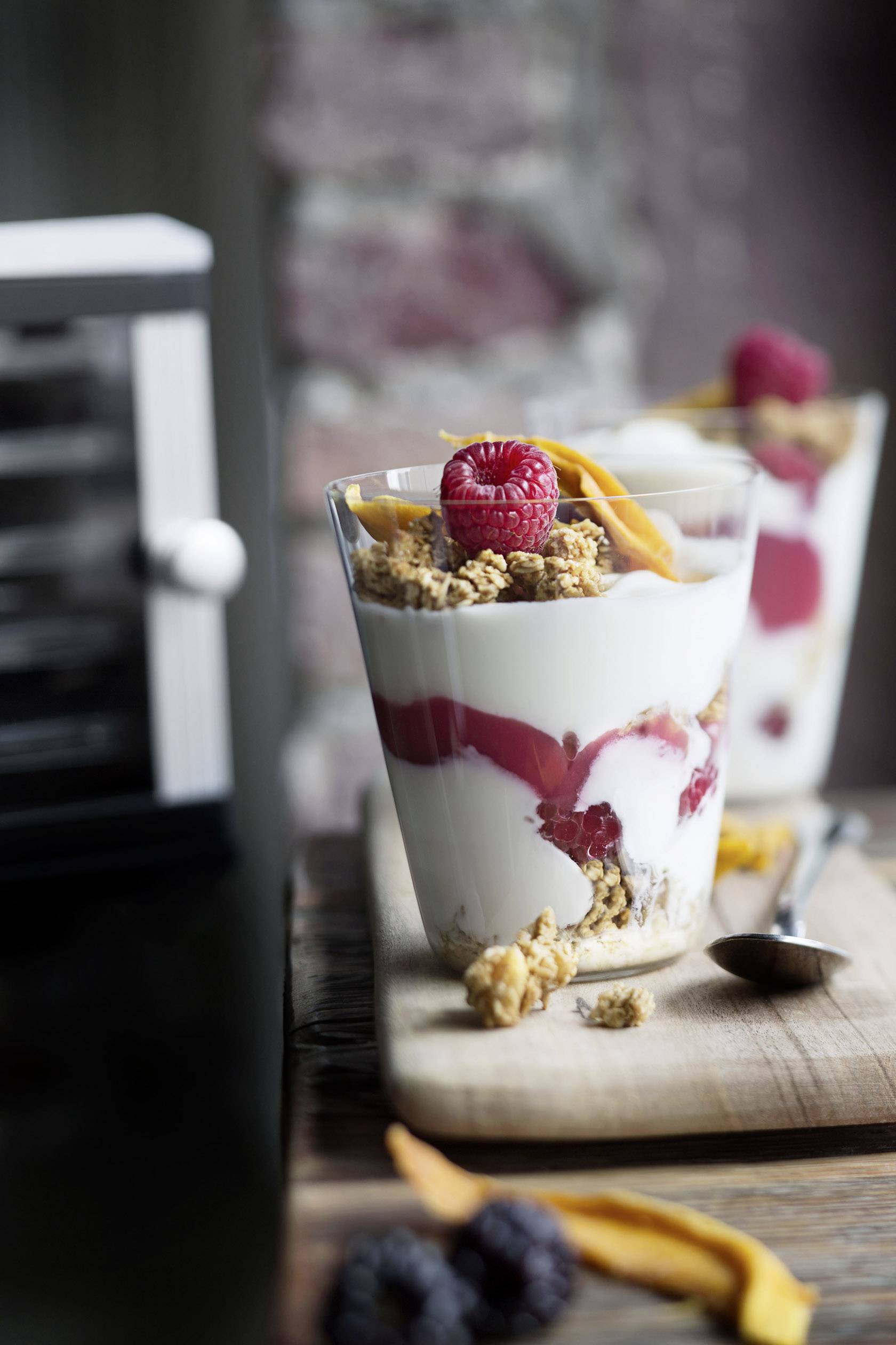 Un bicchiere di dessert a strati con yogurt, lamponi, muesli e strisce di mango essiccato, su un tagliere di legno accanto a un cucchiaio.