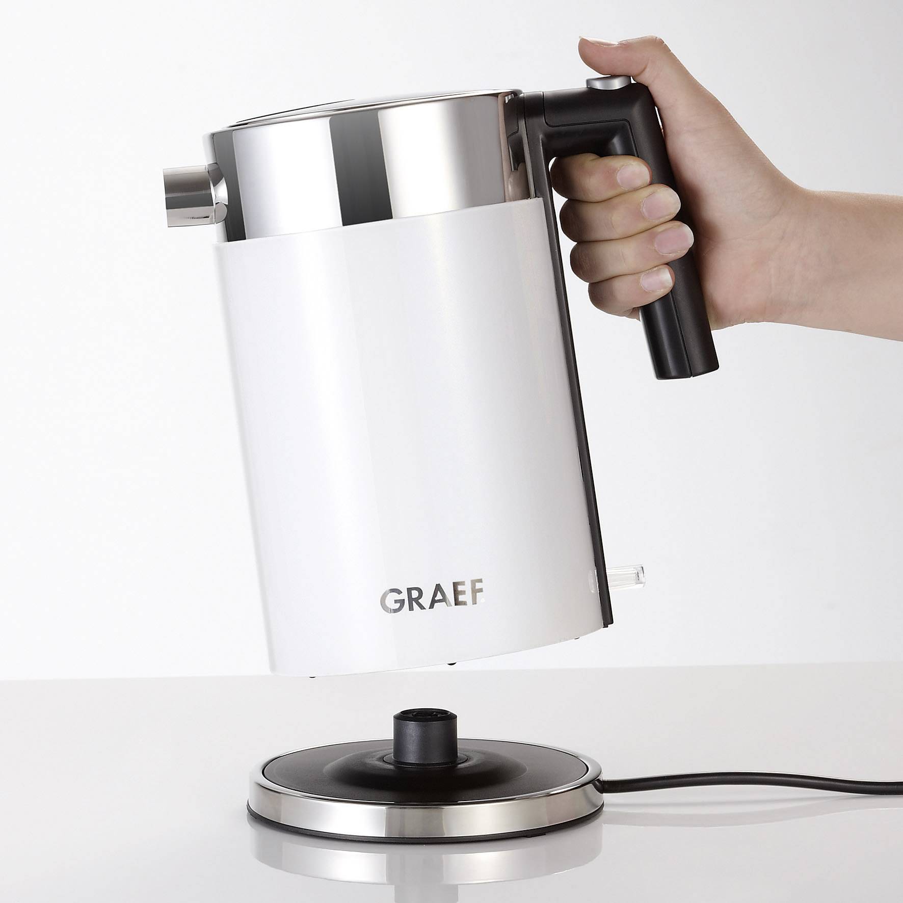 Bollitore elettrico con design moderno sulla stazione di riscaldamento, sorretto da una mano che ne afferra il manico. Nome del marchio 'GRAEF' sulla parte anteriore.