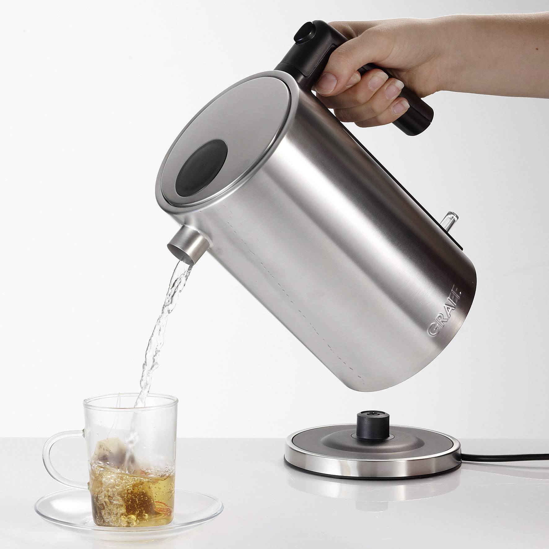 Una mano versa acqua da un bollitore in acciaio inossidabile in una tazza con una bustina di tè.