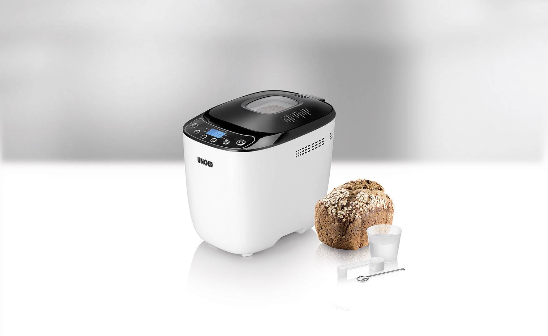 Una macchina per il pane bianca con un display digitale, accanto a un pane appena sfornato, un misurino e un cucchiaio dosatore.