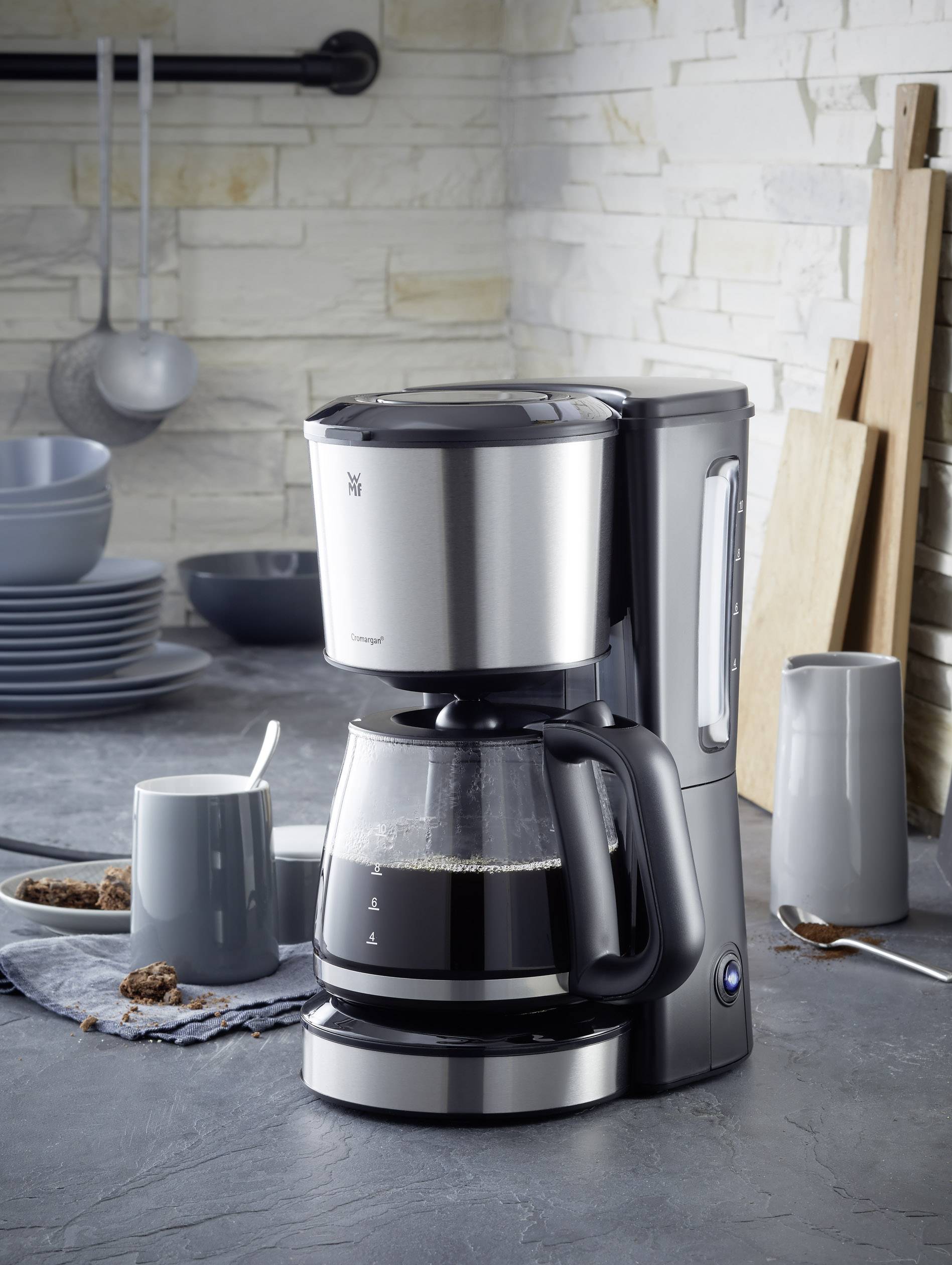Macchina da caffè sul bancone della cucina, circondata da tazze da caffè e dolcetti, davanti a un muro di pietra chiaro.