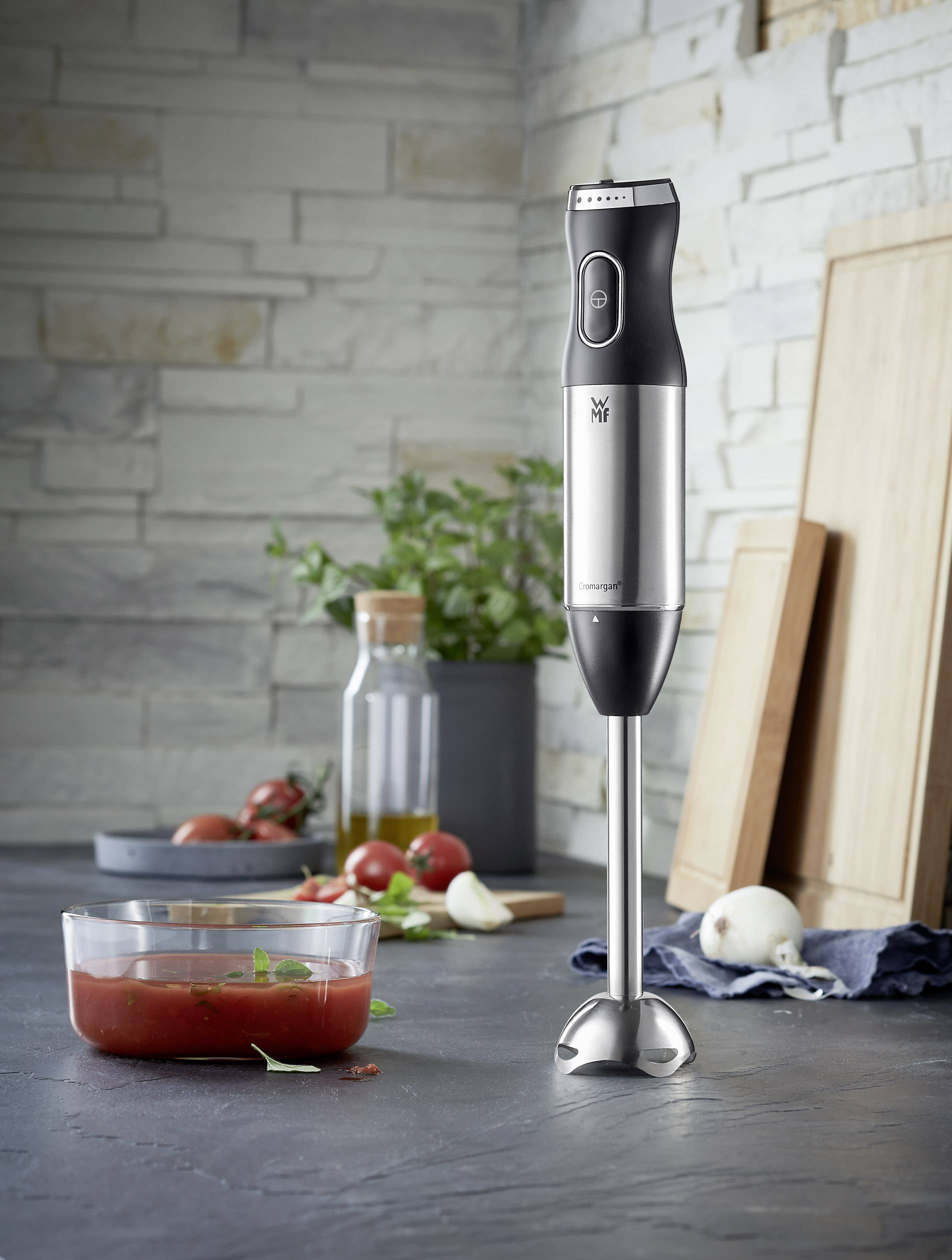 Un frullatore a immersione è posizionato su un piano da cucina accanto a una ciotola di sugo di pomodoro e verdure fresche. Sullo sfondo si vedono utensili da cucina e piante.
