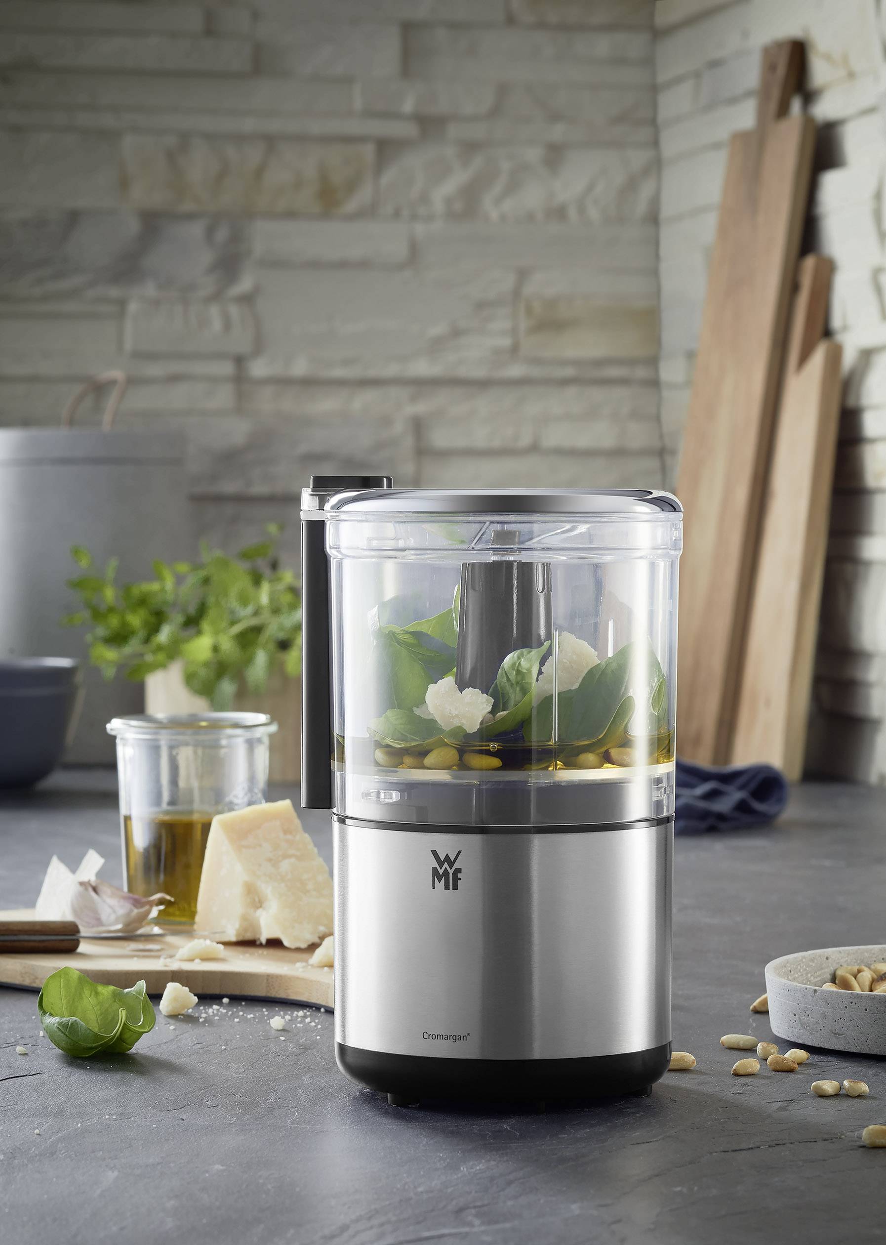 Mixer da cucina argentato su una superficie grigia, riempito con basilico e pezzetti di Parmigiano. Sullo sfondo erbe aromatiche e olio d'oliva.