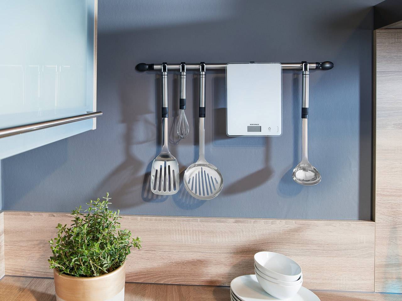 Utensili da cucina e una bilancia sono appesi a una barra in una cucina moderna. Accanto, un vaso con erbe è posizionato sul piano di lavoro.