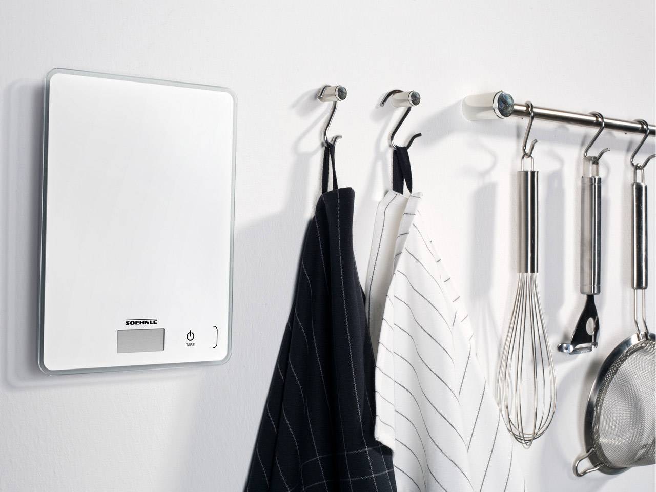 Bilancia digitale da cucina appesa al muro, accanto a cui sono appesi strofinacci e utensili da cucina su ganci.