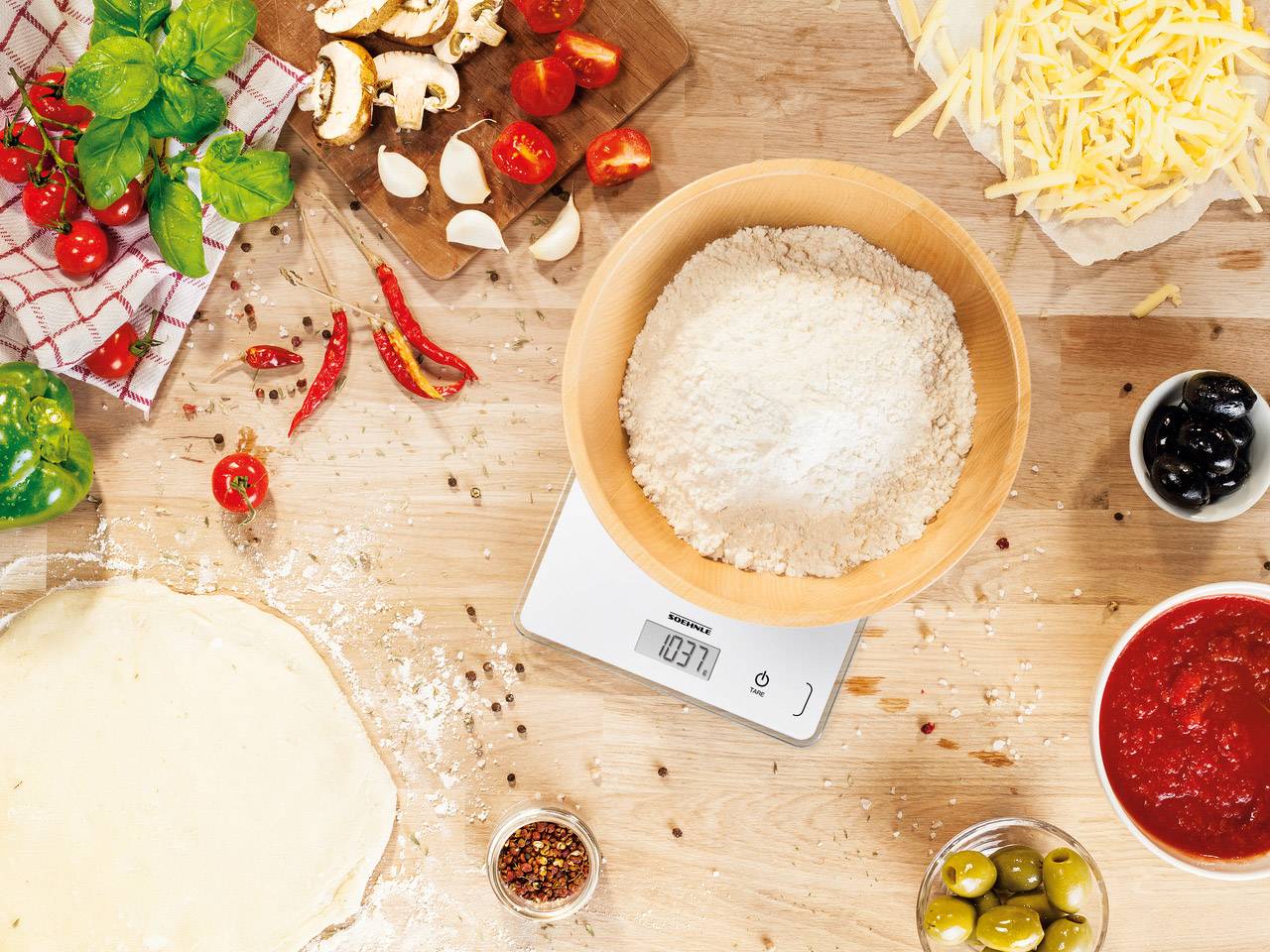 Ingredienti su tagliere di legno con bilancia digitale: farina in ciotola, salsa di pomodoro, formaggio grattugiato, pomodori, funghi, olive. Preparazione della pizza.