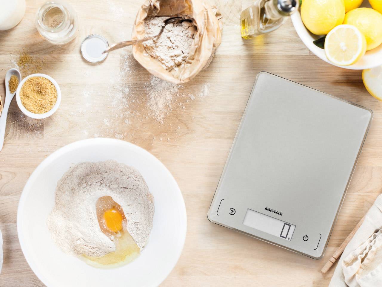 Bilancia da cucina su un tavolo con farina, uova, zucchero e limoni, che indicano la preparazione di una ricetta da forno.