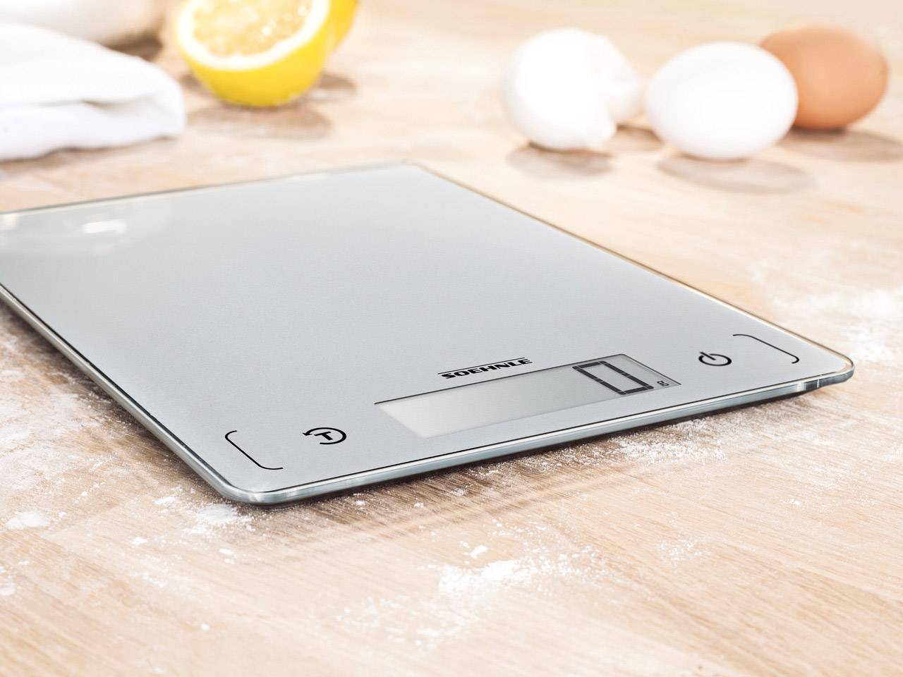 Una bilancia digitale da cucina è posizionata su un tavolo di legno, circondato da farina, uova e metà di un limone sullo sfondo.