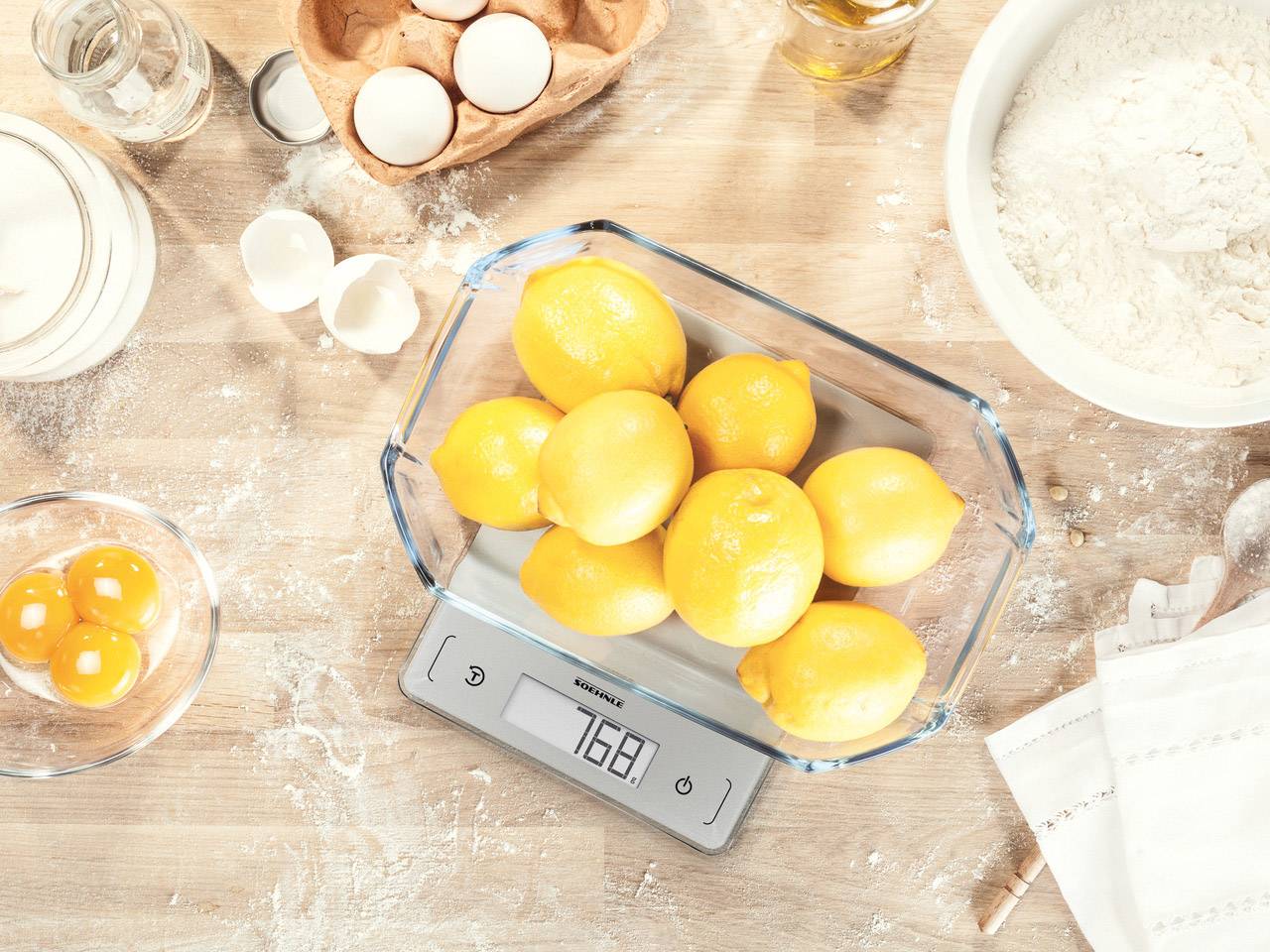Limoni in una ciotola di vetro su una bilancia digitale da cucina che indica 768 grammi, circondati da ingredienti per la preparazione di dolci.
