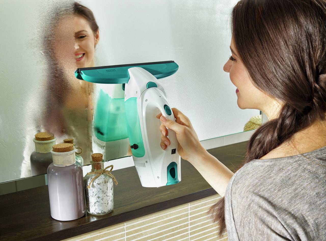 Una donna sta pulendo uno specchio del bagno con un detergente elettrico per vetri. Due bicchieri con prodotti per il bagno sono posizionati sul lavandino.