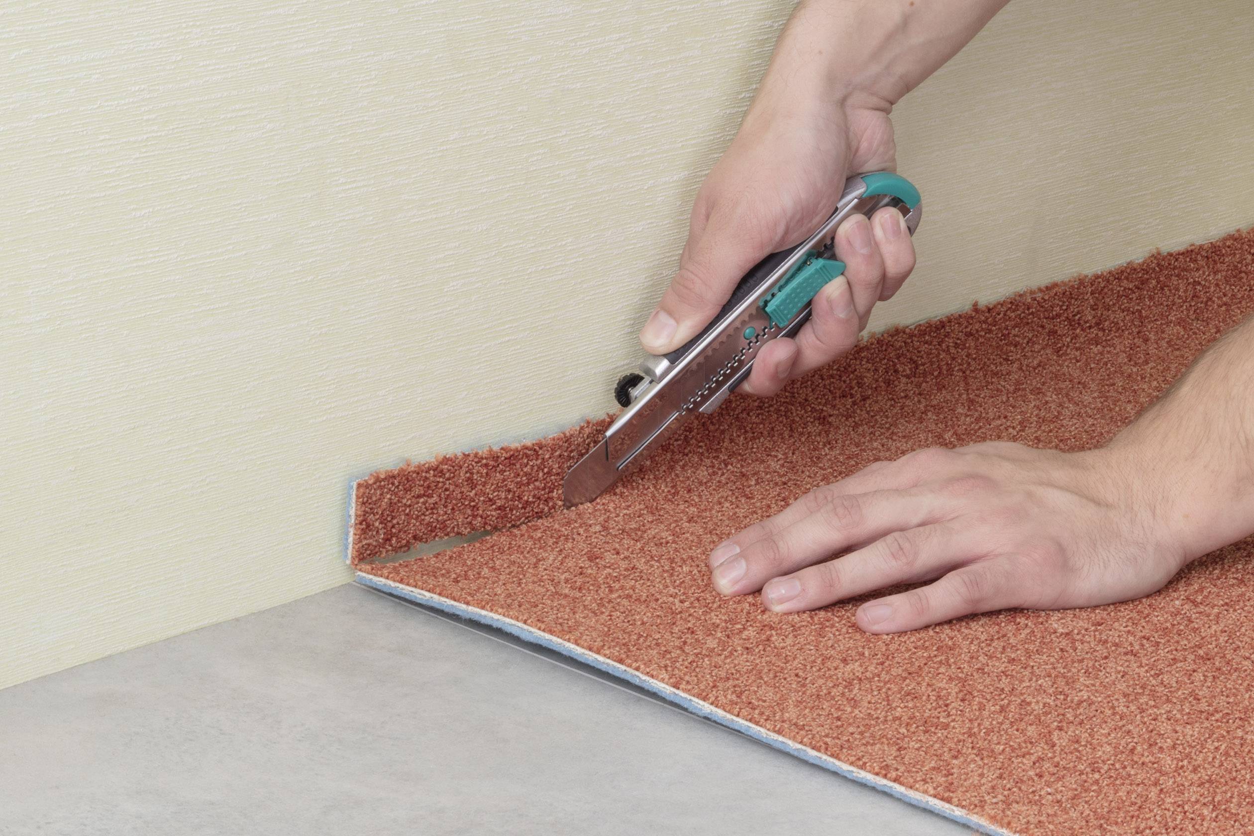 Una persona taglia un tappeto rosso-marrone lungo un bordo utilizzando un taglierino. Il tappeto è posizionato su un rivestimento per pavimenti.