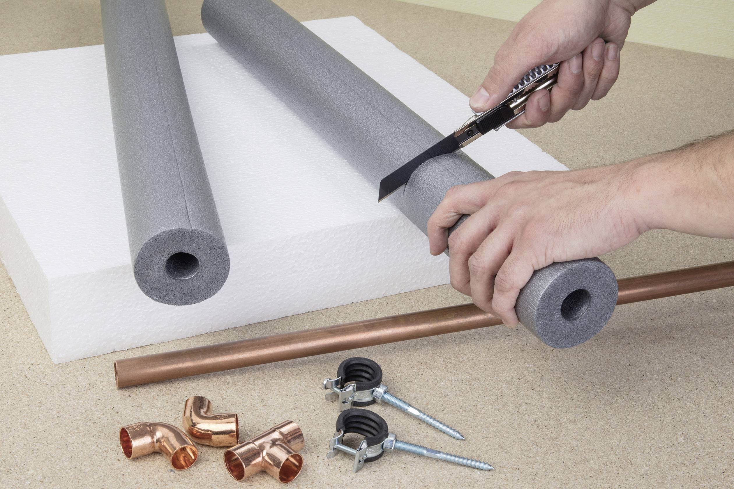 Due mani tagliano un isolamento grigio per tubazioni con un cutter; un tubo di rame e accessori sono posizionati su un tavolo.
