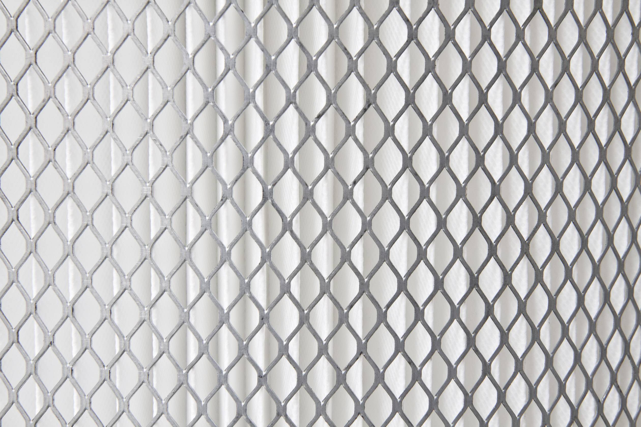 Griglia metallica con motivo a rombi che lascia passare la luce e proietta ombre su una superficie chiara sottostante.