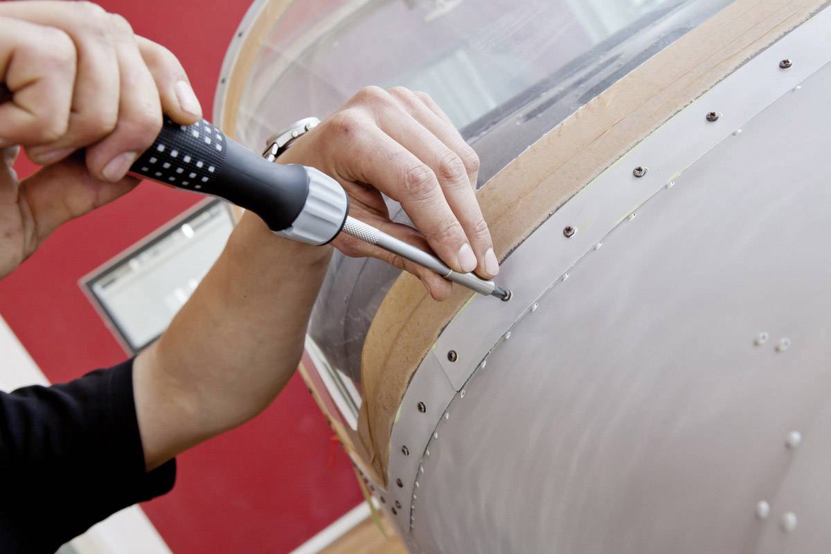Una persona sta serrando delle viti su una fusoliera di aereo. Il primo piano mostra le mani al lavoro con un cacciavite.