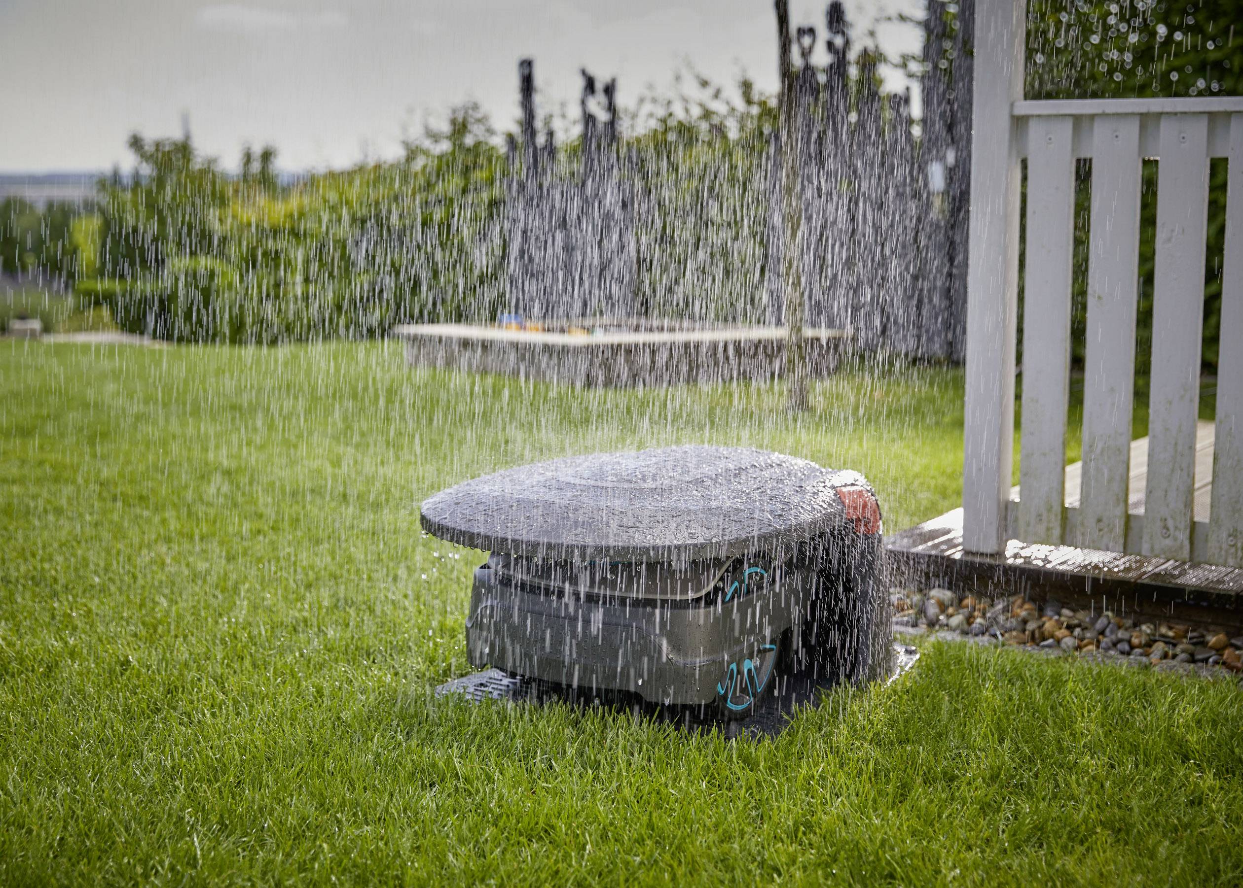 Un irrigatore spruzza acqua su un giardino verde. Un recinto bianco è visibile sullo sfondo. L'acqua forma una pioggia uniforme.