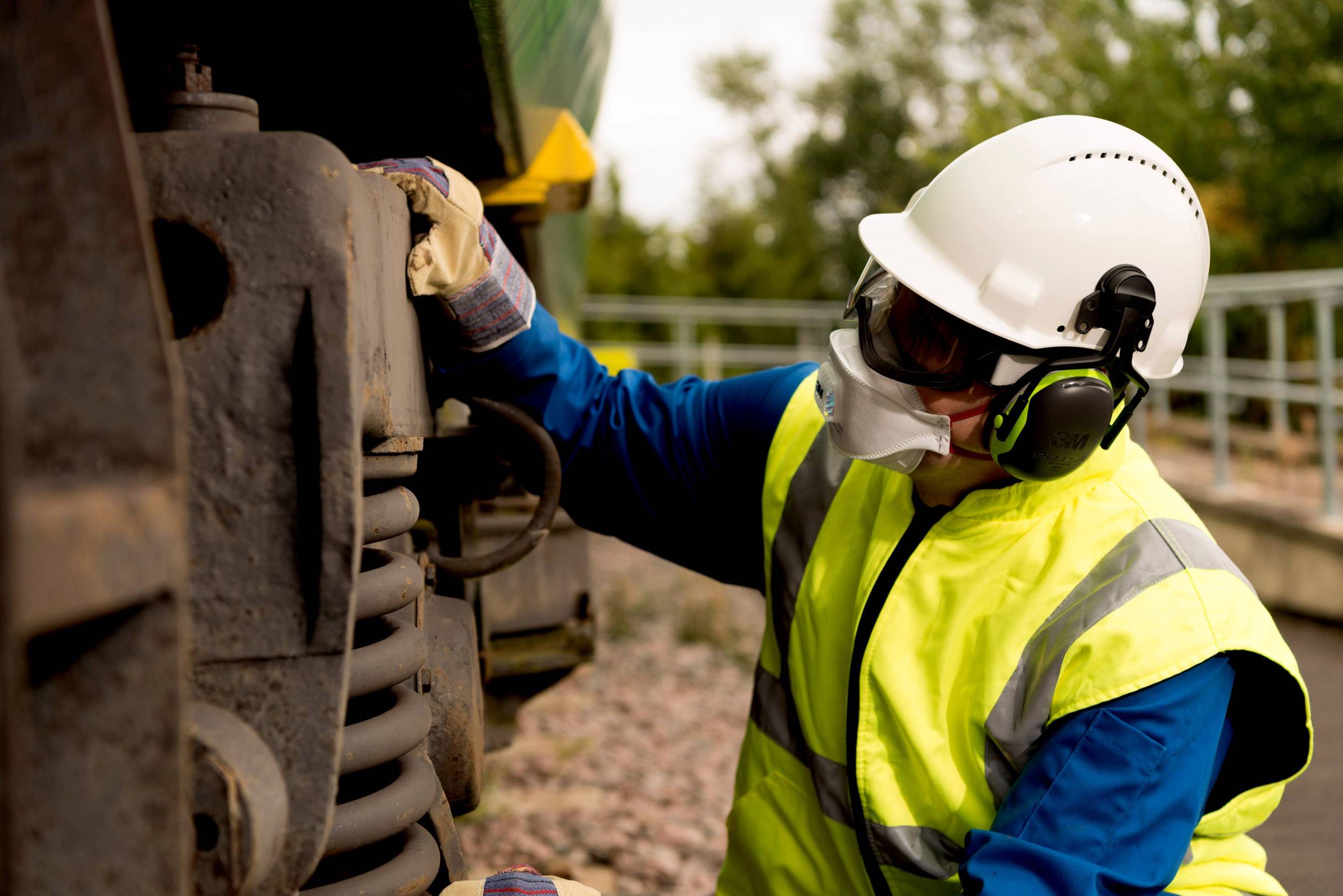 Un lavoratore indossa dispositivi di protezione individuale, un elmetto e protezioni per l'udito. Sta ispezionando o effettuando manutenzione ai componenti meccanici di un treno all'aperto.