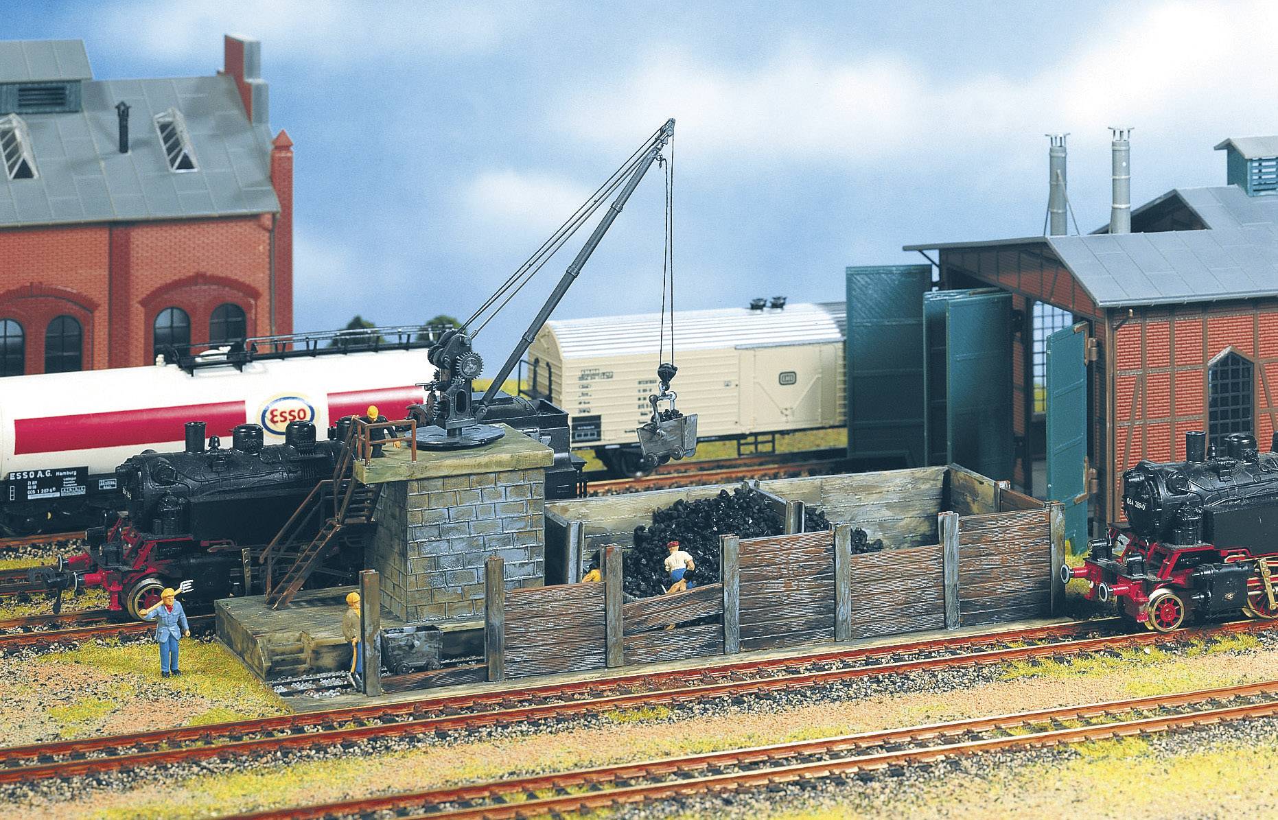 Una scena ferroviaria in miniatura mostra una stazione merci con locomotive a vapore, un deposito di carbone e un punto di carico. Una gru è pronta per le operazioni.