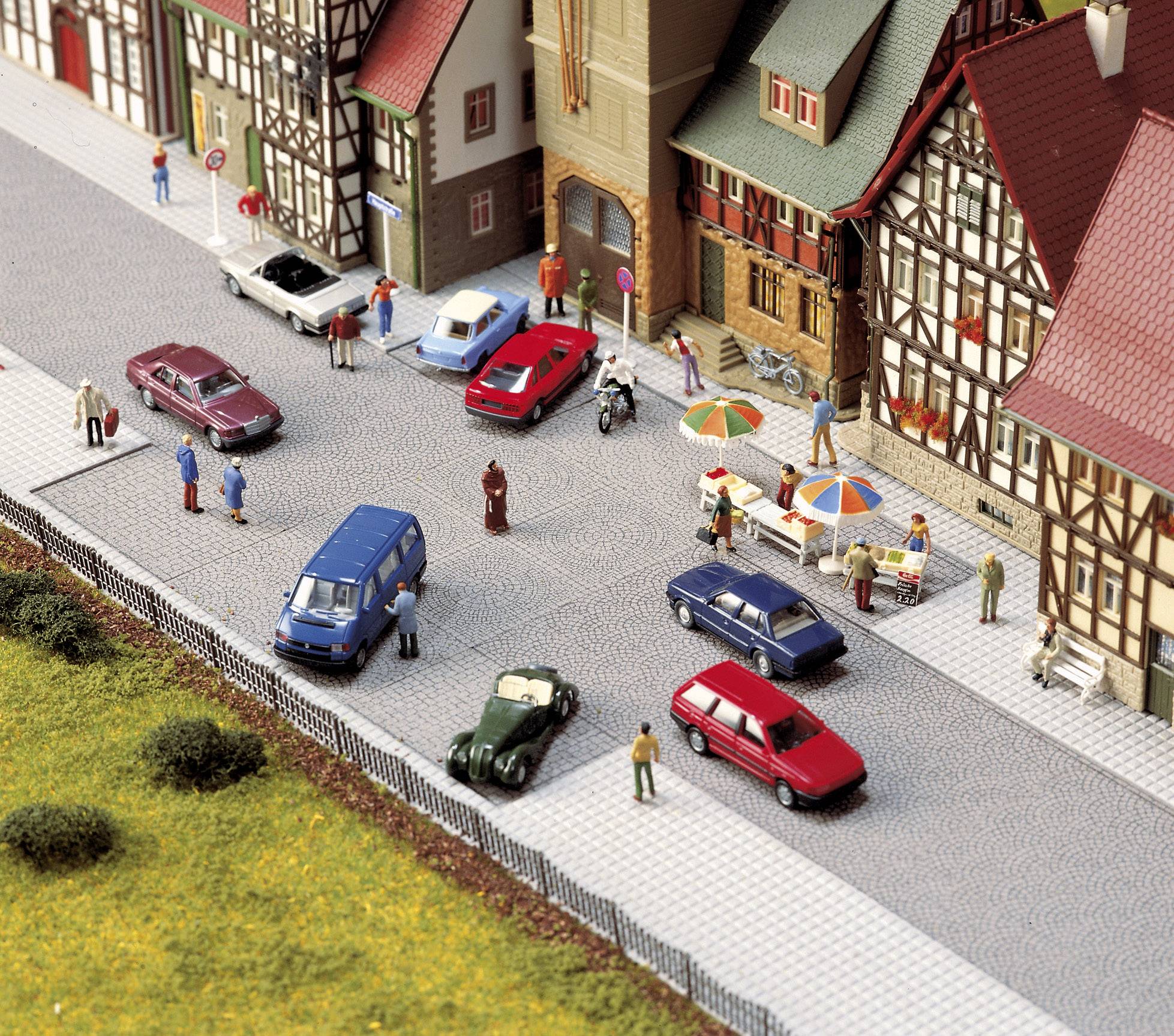 Scena di strada in una città in miniatura con case a graticcio, diverse auto, pedoni e bancarelle. Persone che interagiscono in un ambiente urbano.