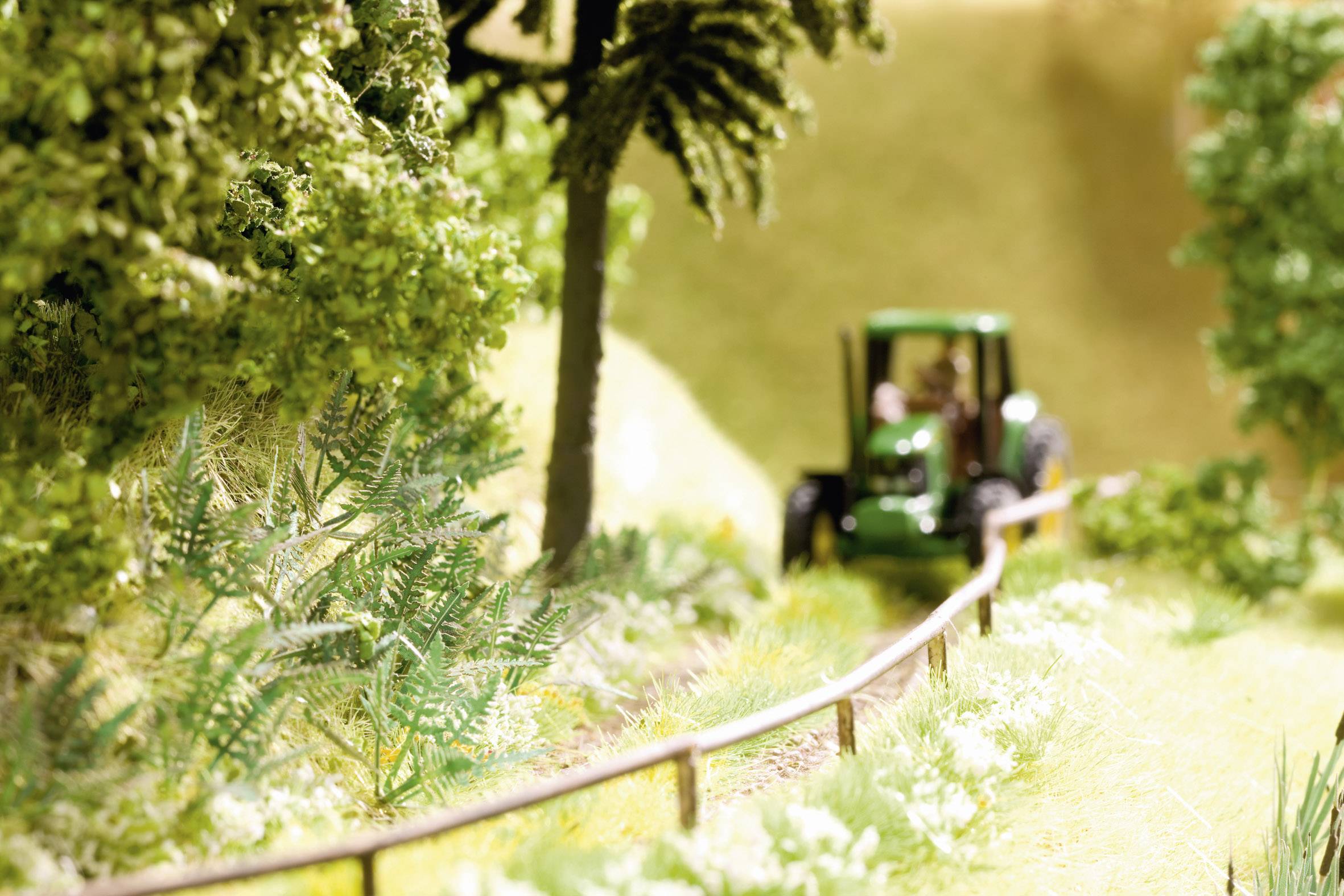 Un piccolo modellino giocattolo di un trattore verde sta guidando su un sentiero di campagna attraverso un rigoglioso prato verde e accanto ad alcuni alberi.