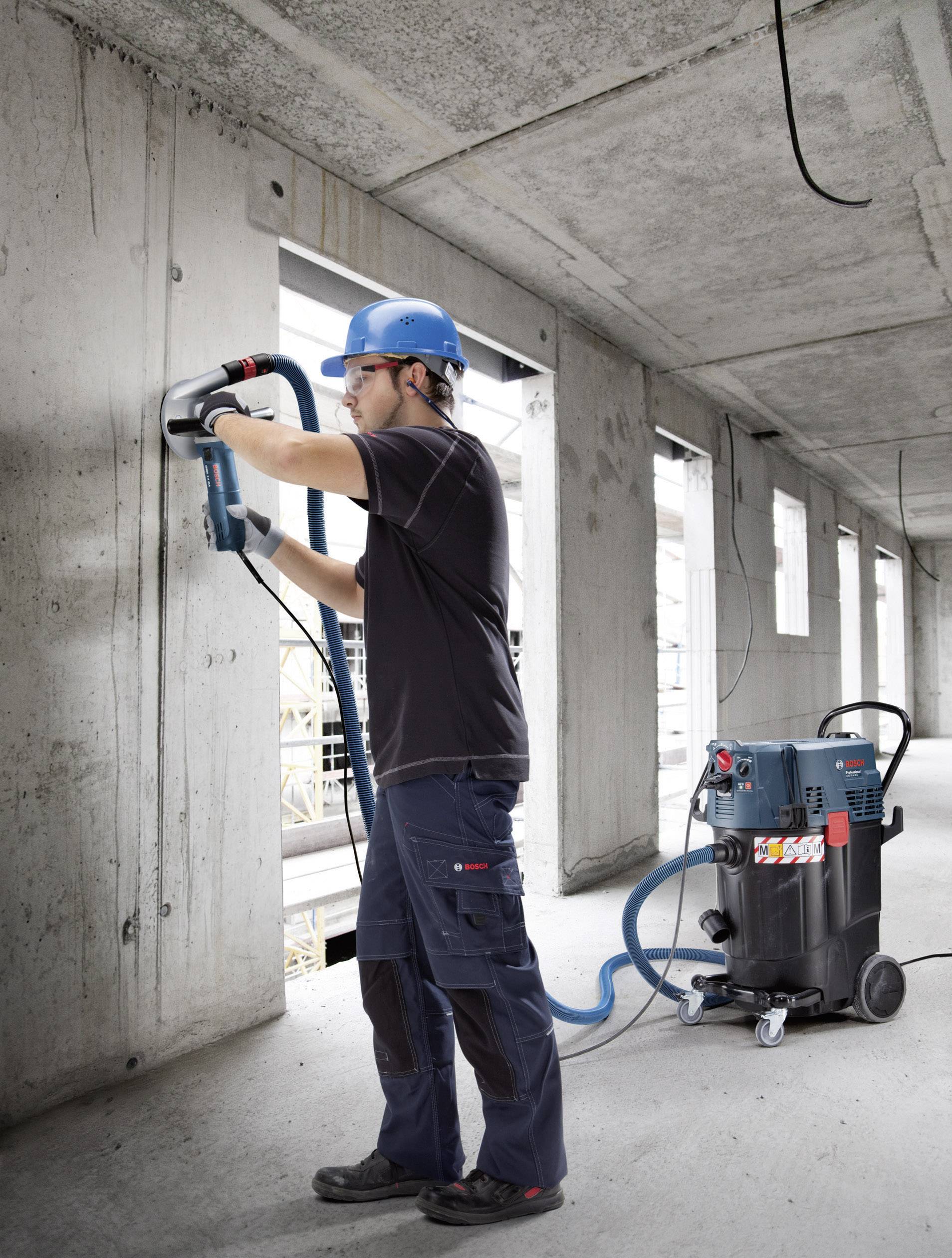 Una persona con un casco protettivo sta lavorando con un martello pneumatico su una parete in calcestruzzo in un edificio parzialmente completato. Un aspiratore industriale è posizionato accanto a lui.