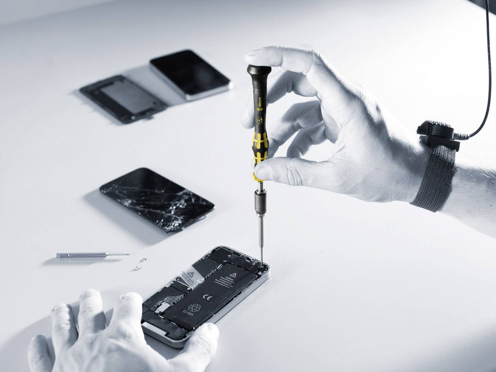 Una persona sta riparando uno smartphone con un cacciavite. Sullo sfondo sono posizionati altri dispositivi e strumenti.