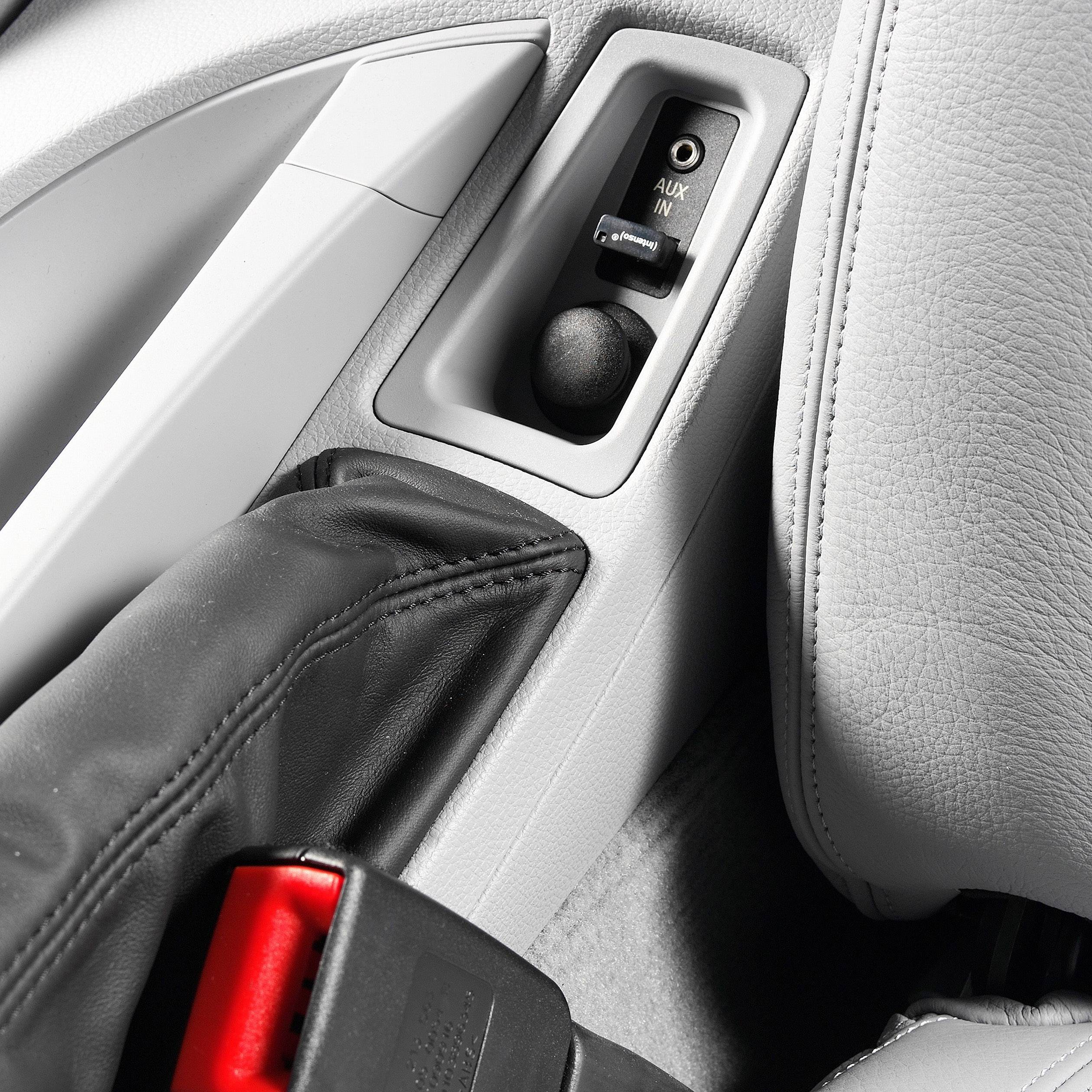 Interno di un'auto che mostra un ingresso AUX tra sedili in pelle, accanto a una cintura di sicurezza, idoneo per connessioni musicali.