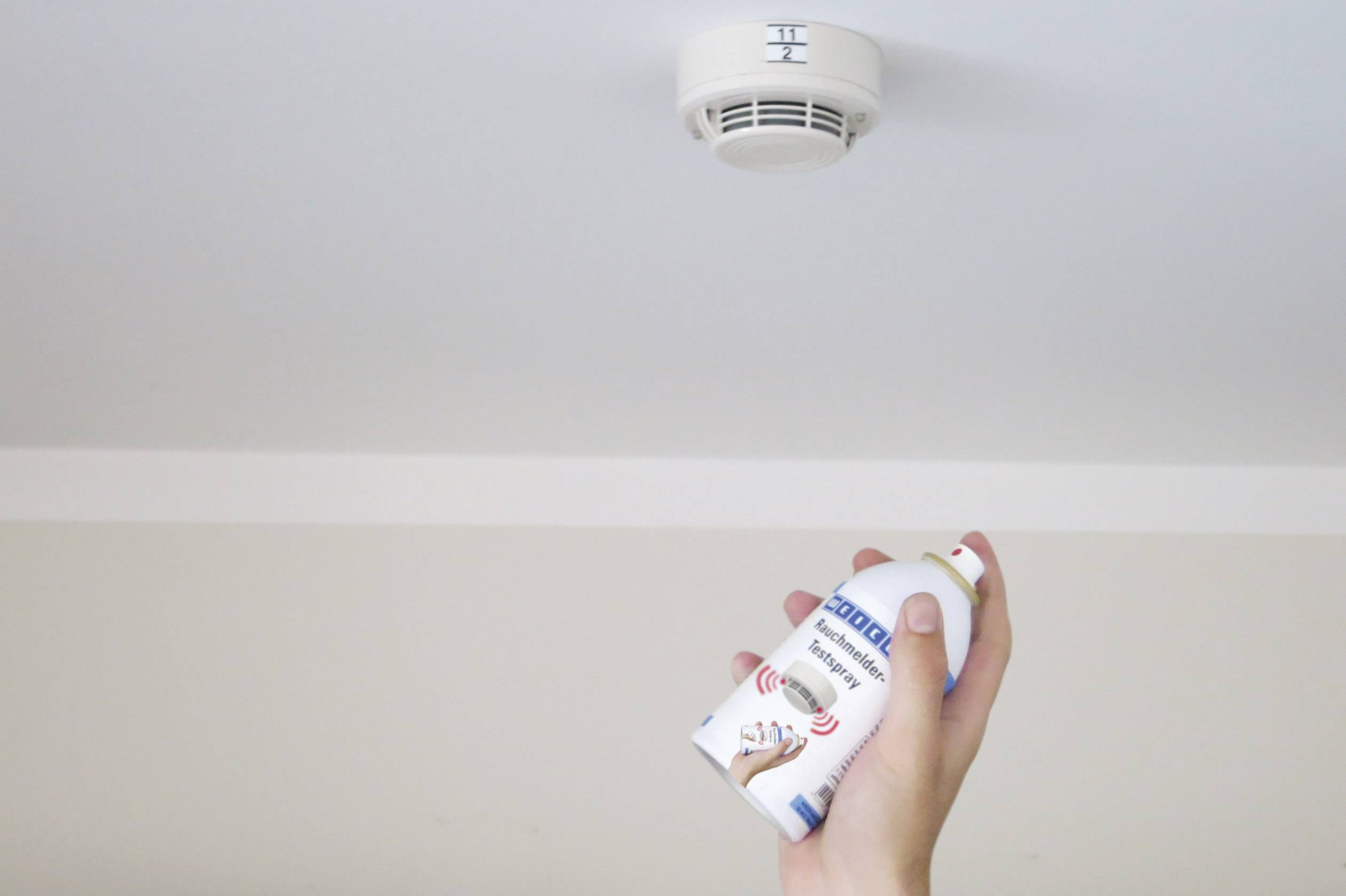 Una mano sta testando un rilevatore di fumo sul soffitto con uno spray per verificare il corretto funzionamento del dispositivo.