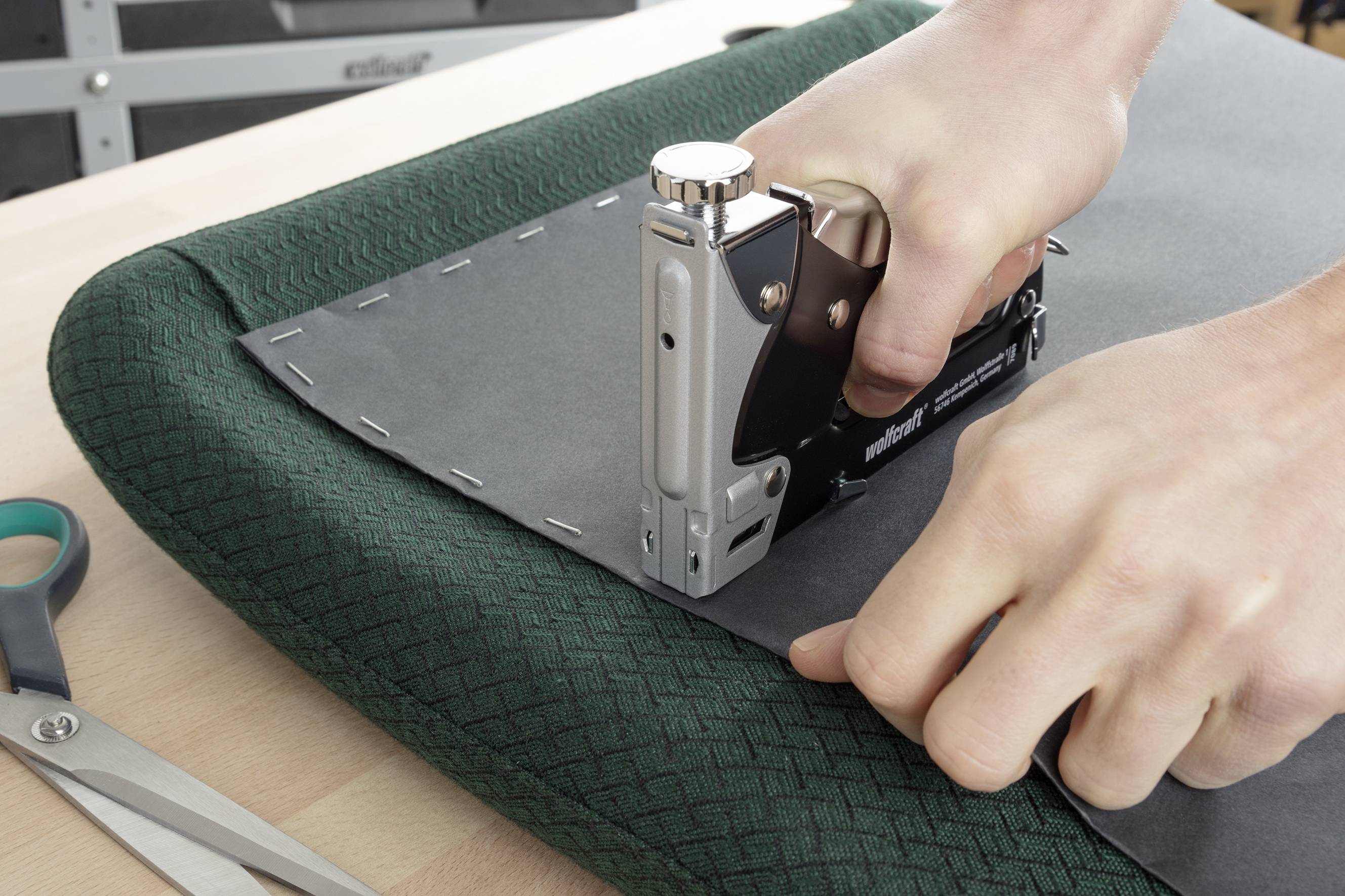 Una persona fissa il tessuto con una pistola sparapunti su un'imbottitura. Il tessuto è grigio scuro e il rivestimento dell'imbottitura è verde a motivi. Le forbici sono poggiate accanto.
