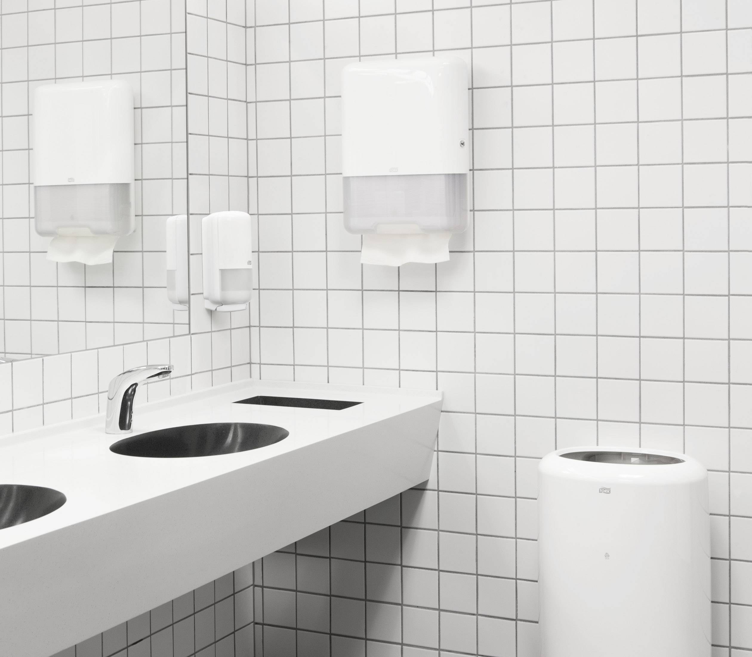 Due lavandini con rubinetti in un bagno pubblico bianco e piastrellato; sulla parete sono montati dei dispenser di sapone.