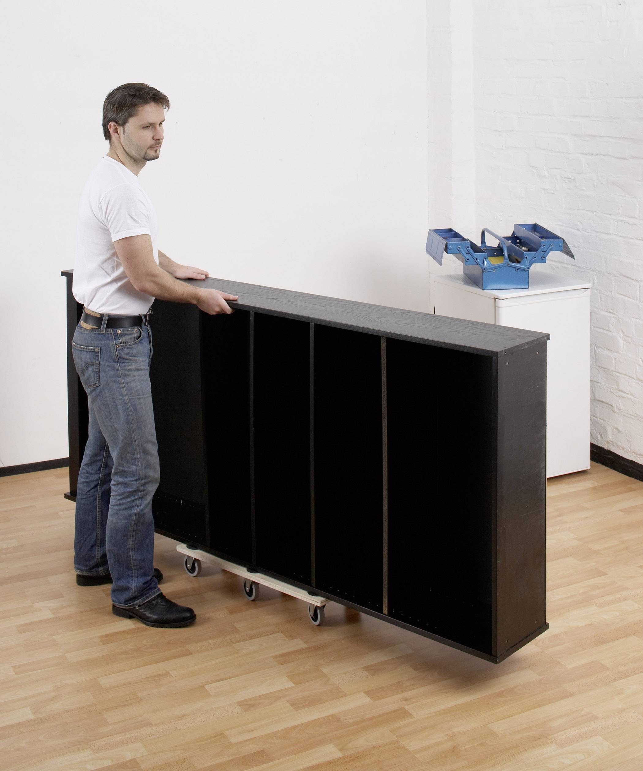 Una persona sta spingendo una grande libreria nera su ruote in una stanza con pavimento in legno. Sullo sfondo, c'è un tavolo bianco.