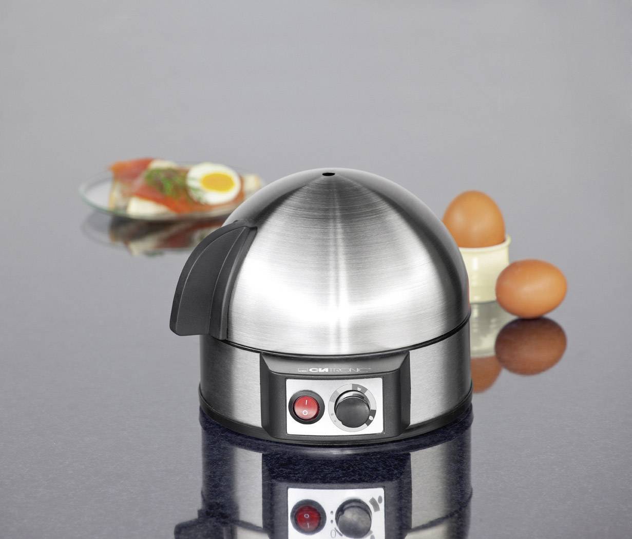 Un cuocuova in acciaio inossidabile su una superficie liscia, con due uova accanto. Sullo sfondo un piatto con un panino farcito.