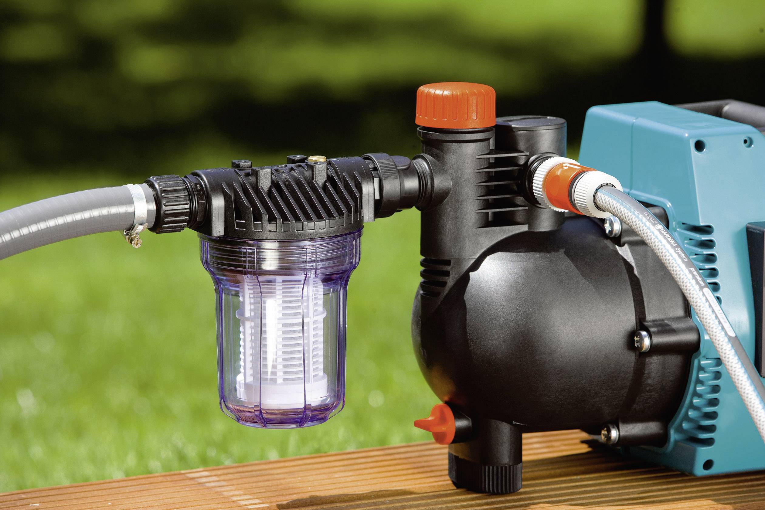 Una pompa da giardino con camera di filtraggio trasparente e attacchi per tubi che aspira acqua da un tavolo di legno; sullo sfondo erba.