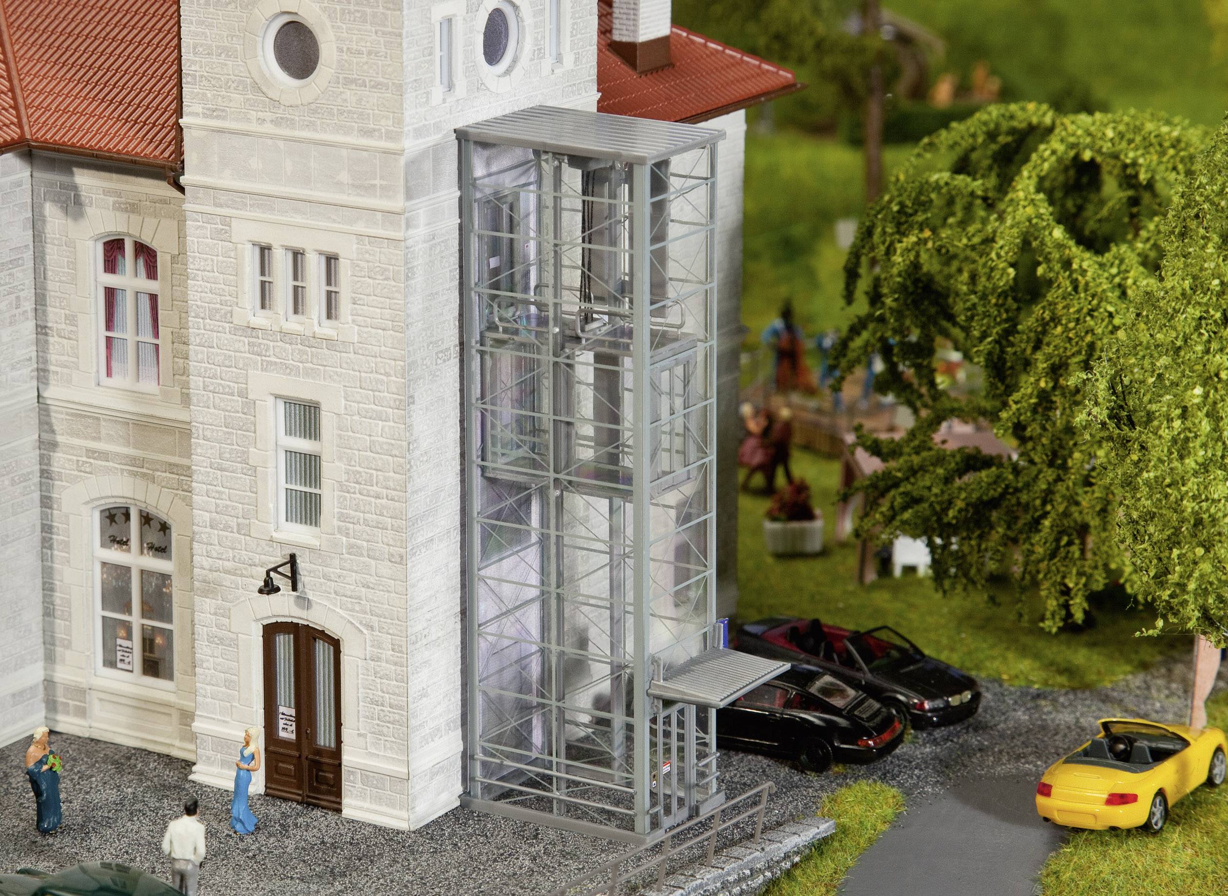 Un edificio in miniatura con un ascensore di vetro, circondato da alberi e auto parcheggiate. Persone sono in piedi e chiacchierano davanti all'edificio.