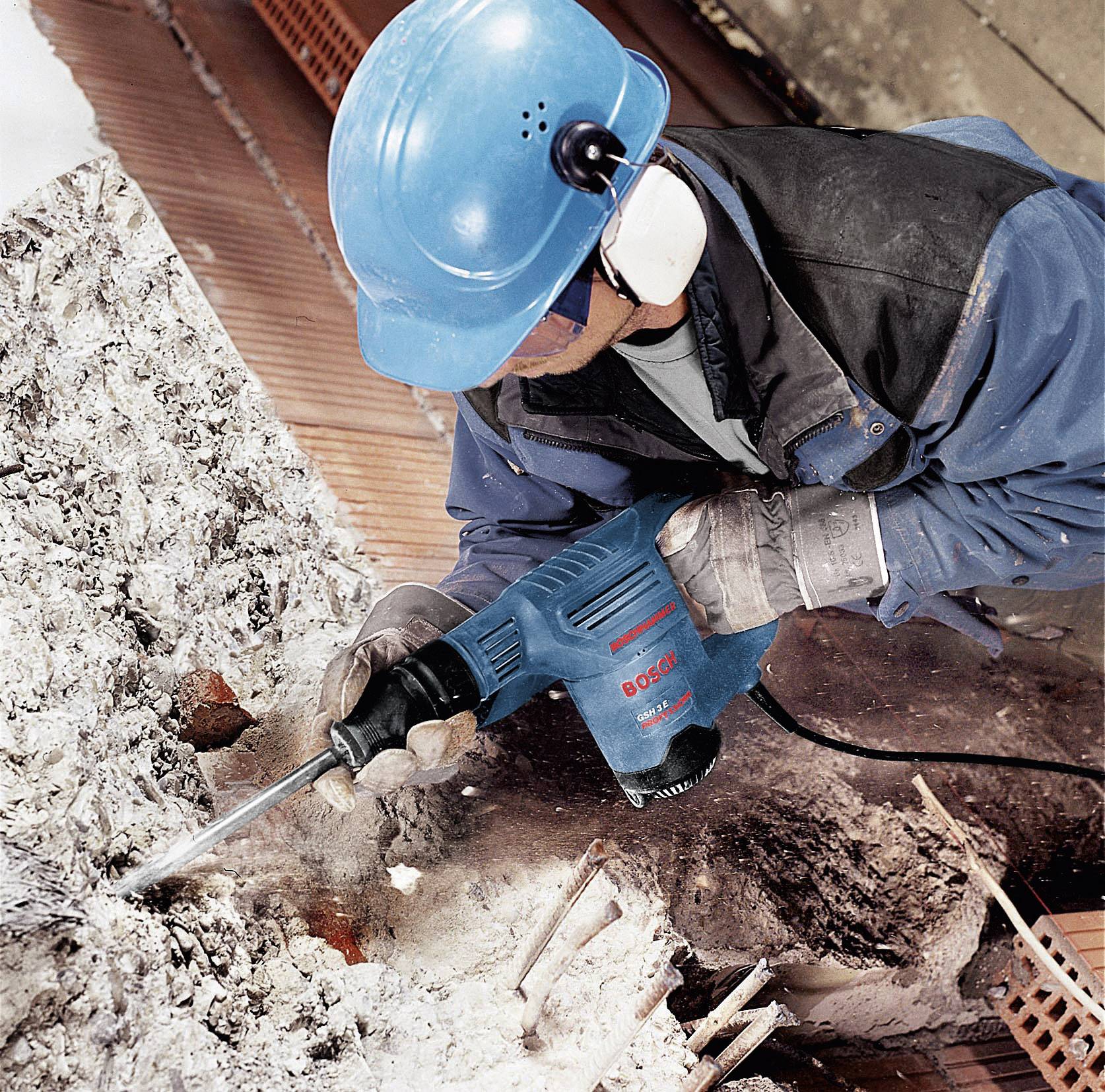 Una persona con casco di protezione e protezioni acustiche sta trapanando detriti di cemento in un cantiere con un trapano blu.
