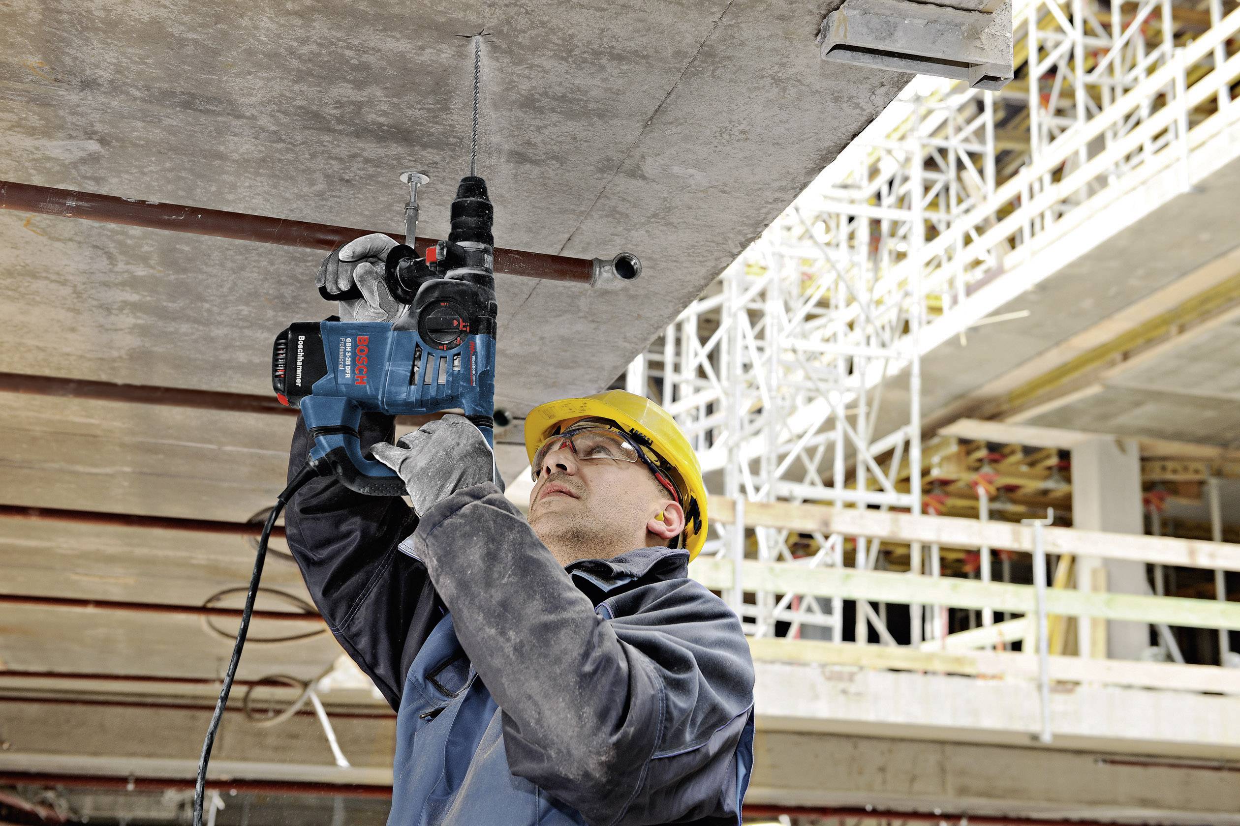 Un operaio edile con indumenti protettivi perfora un soffitto in calcestruzzo dal basso utilizzando un macchinario. Sullo sfondo è visibile un ponteggio.