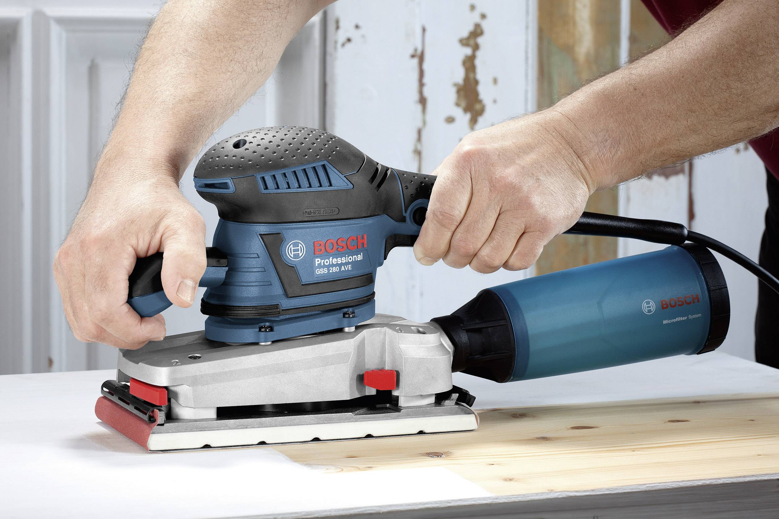 Una persona sta utilizzando un levigatrice orbitale Bosch Professional su una superficie in legno. Sullo sfondo si vedono pannelli di legno invecchiati.