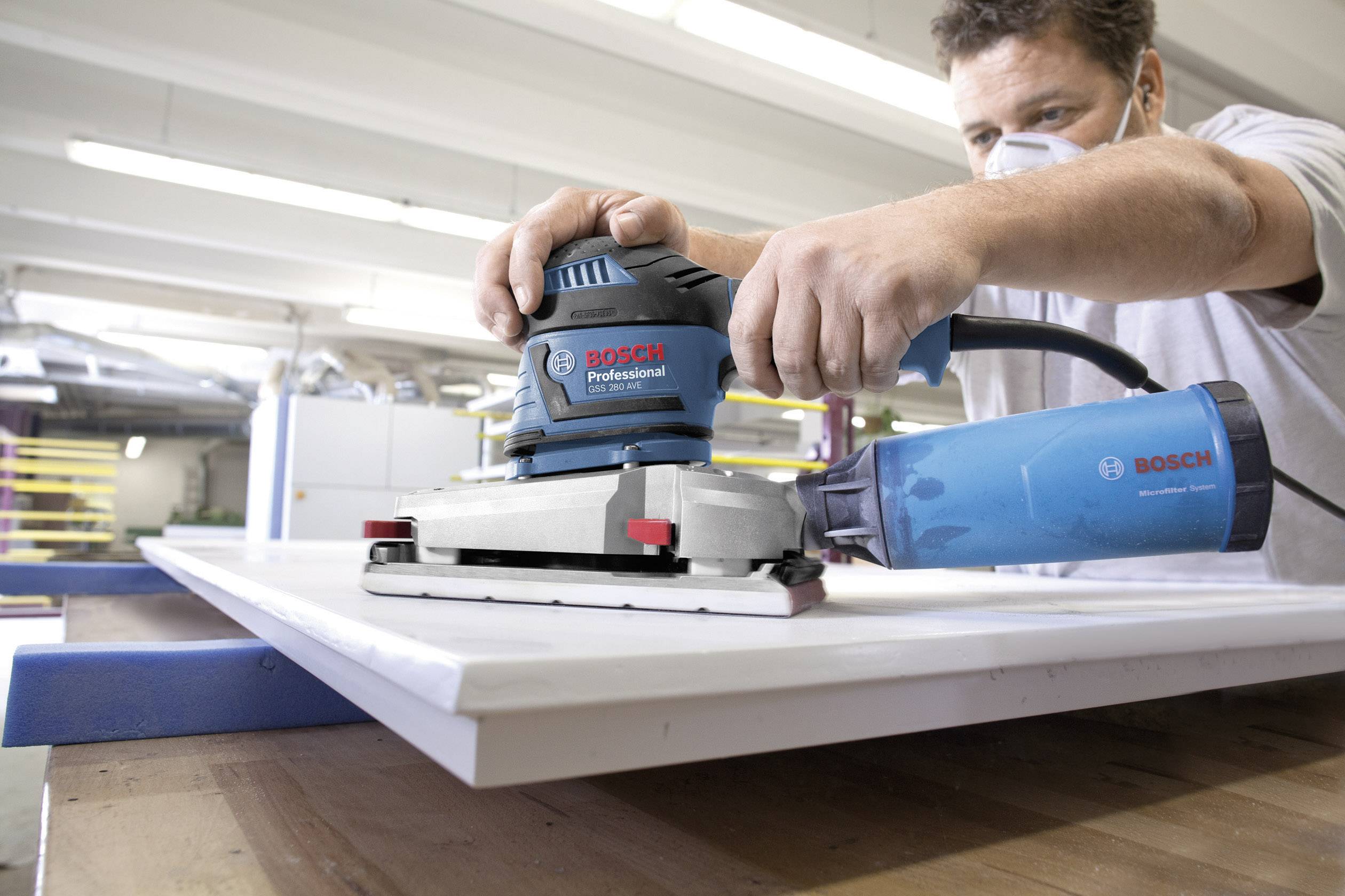 Un uomo con una maschera respiratoria utilizza un levigatrice orbitale Bosch Professional blu per lavorare una tavola di legno bianca in un laboratorio.