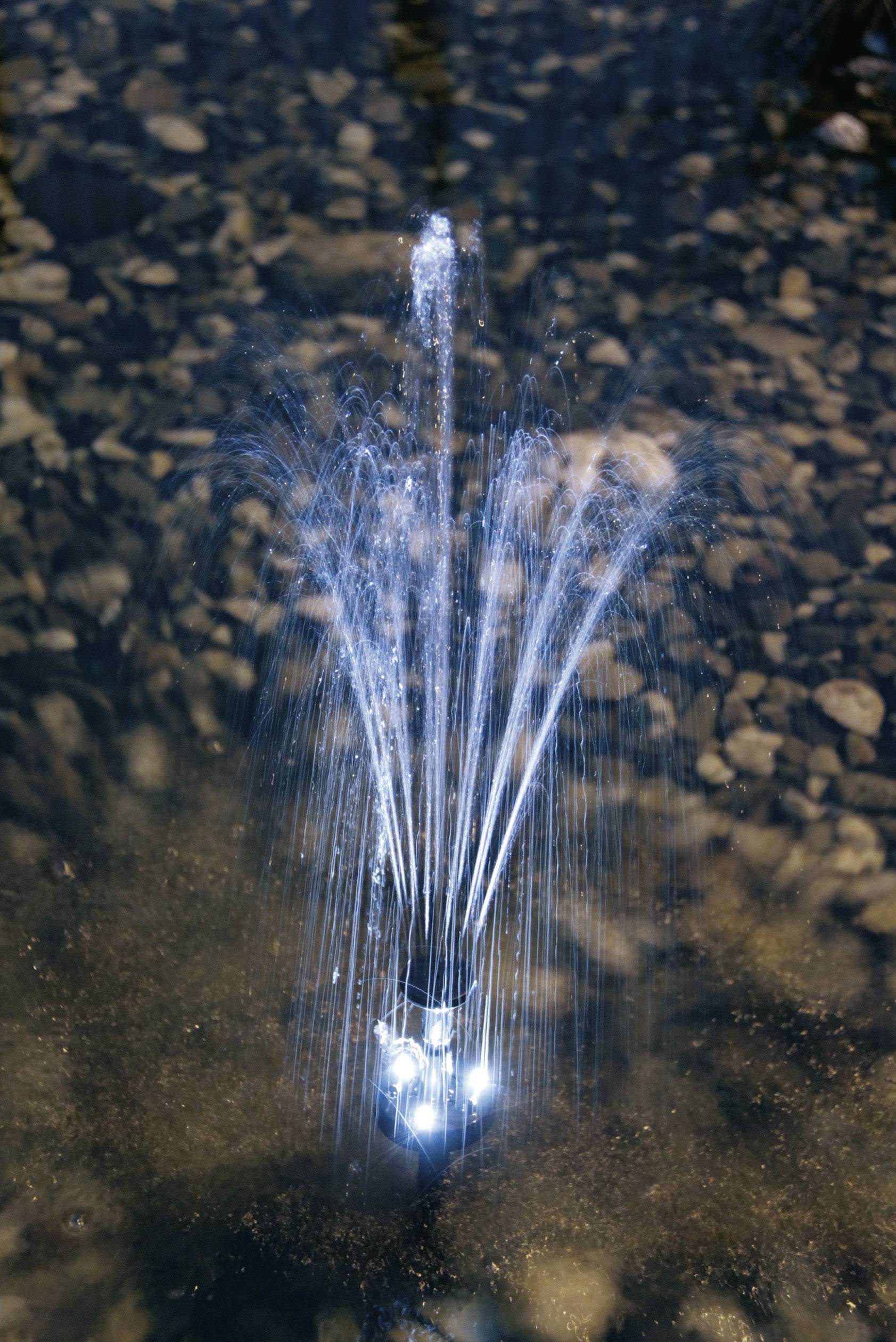 Una fontana illuminata si anima di notte, getti d'acqua formano eleganti archi sopra un terreno sassoso.