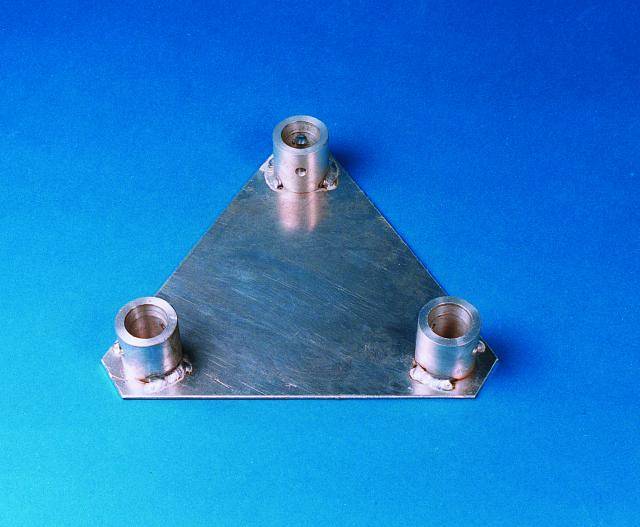 Un pezzo metallico triangolare con tre rilievi cilindrici, poggiato su uno sfondo blu.