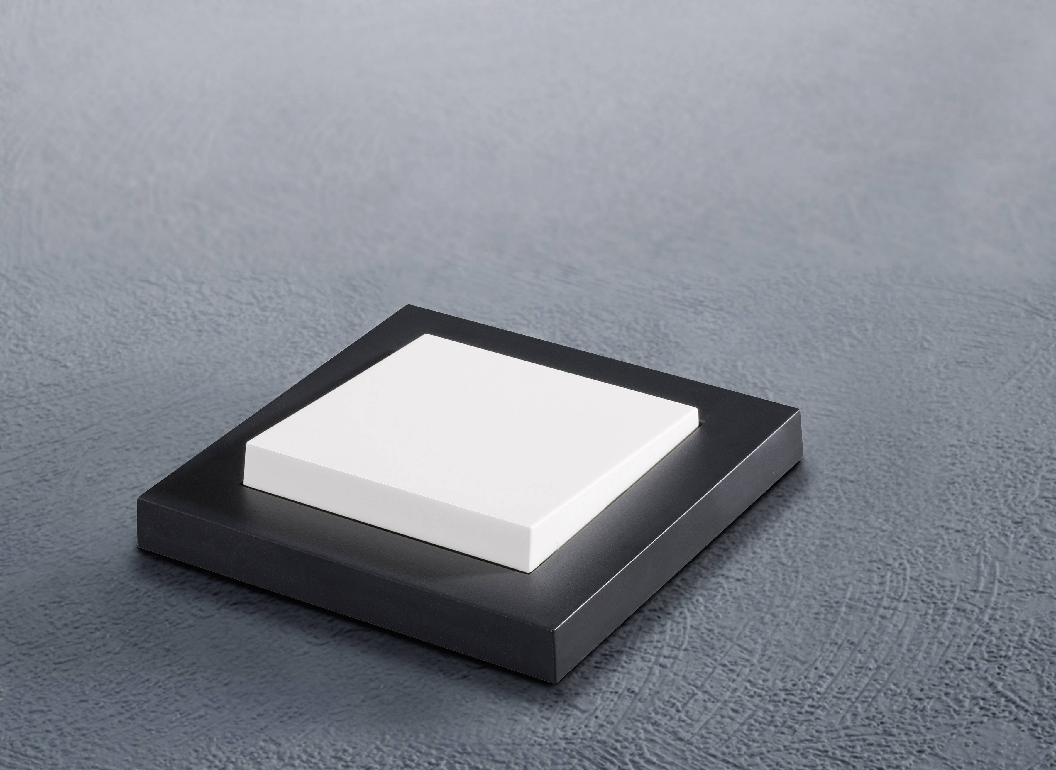 Un oggetto piatto, quadrato, bianco poggia su una base quadrata più grande, nera, su uno sfondo grigio.